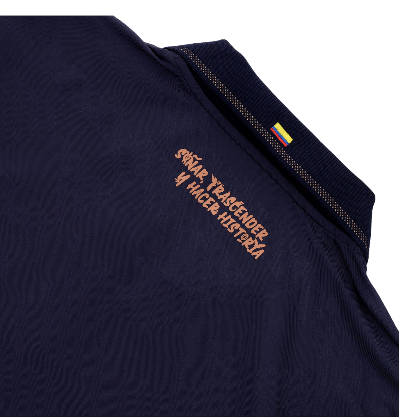A close-up of the back collar area of a dark navy blue shirt. Below the collar, a Spanish slogan is printed in a stylized copper-colored font that reads "SOÑAR, TRASCENDER Y HACER HISTORIA." A small Colombian flag is visible on the ribbed collar, and the entire image is set against a black background.