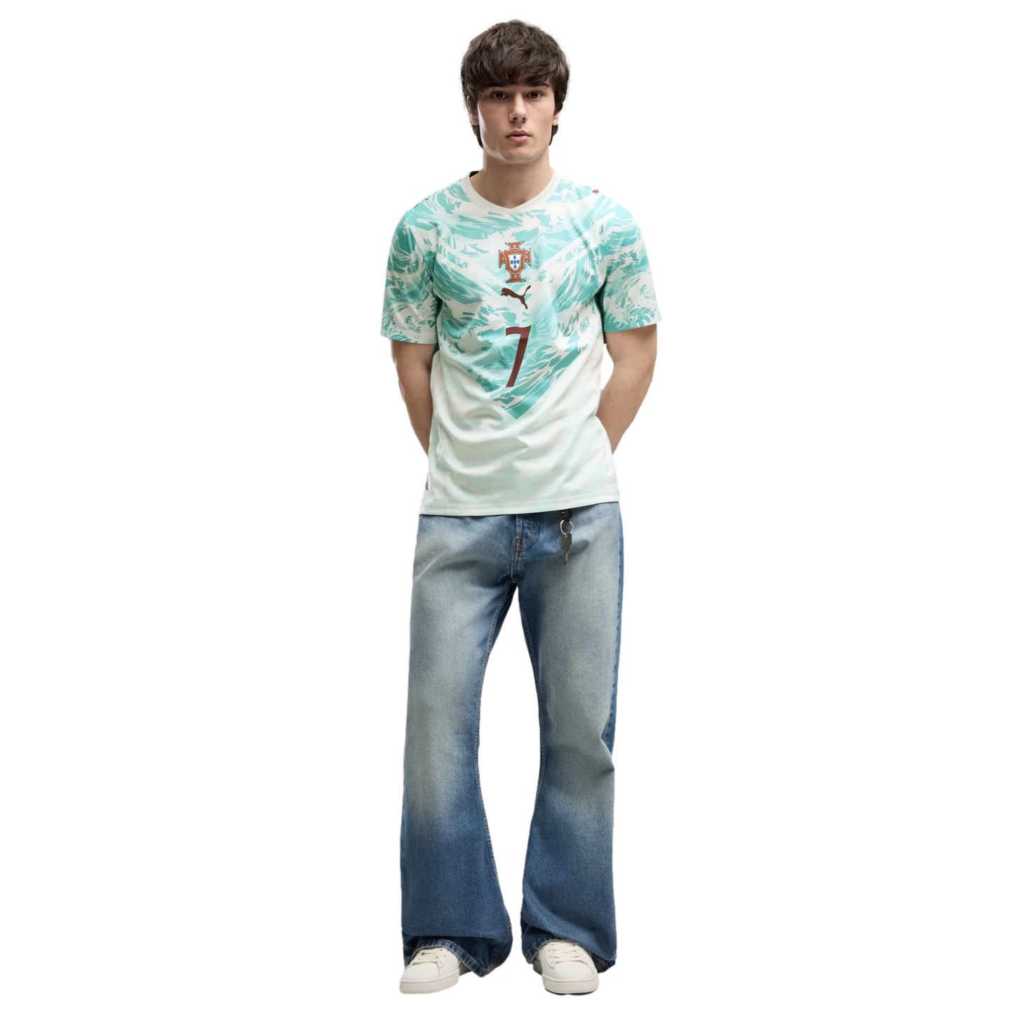 A young man with dark, textured hair stands against a solid black background. He is wearing a light green and teal patterned athletic jersey with a crest and the number 7, wide-leg light-wash blue jeans, and white sneakers. He stands with his hands behind his back, looking directly at the camera.