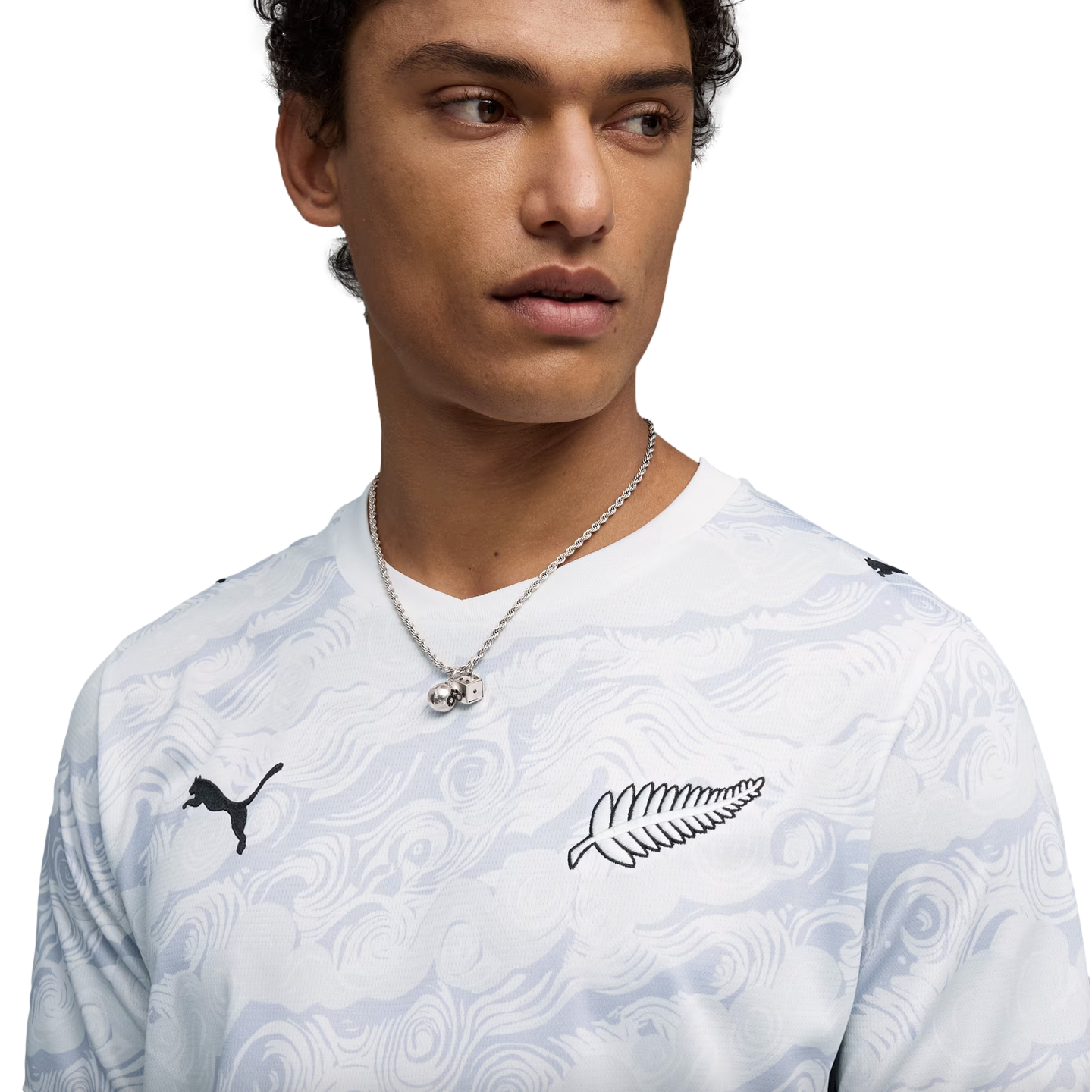 A close-up of a young man wearing a white, wavy-patterned long-sleeve Puma shirt. The shirt features a black Puma logo on the right side and a black silver fern logo on the left. He is also wearing a silver chain necklace with a dice pendant against a solid black background.
