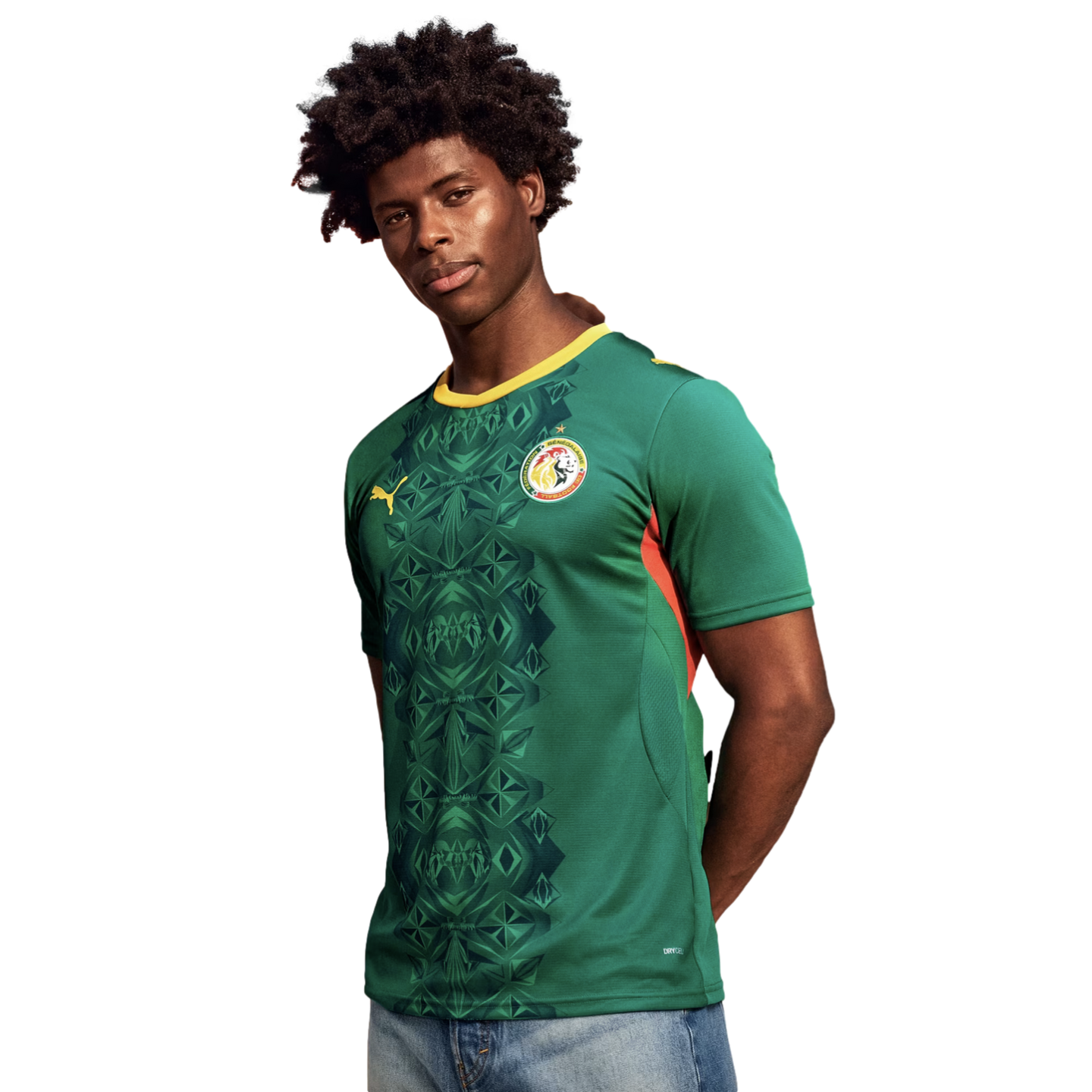 A man with dark, curly hair poses in a green Senegal national team soccer jersey against a solid black background. The jersey features a yellow collar, a dark green geometric pattern down the center, the Puma logo, and the national team crest. He is wearing blue jeans with his arms behind his back.