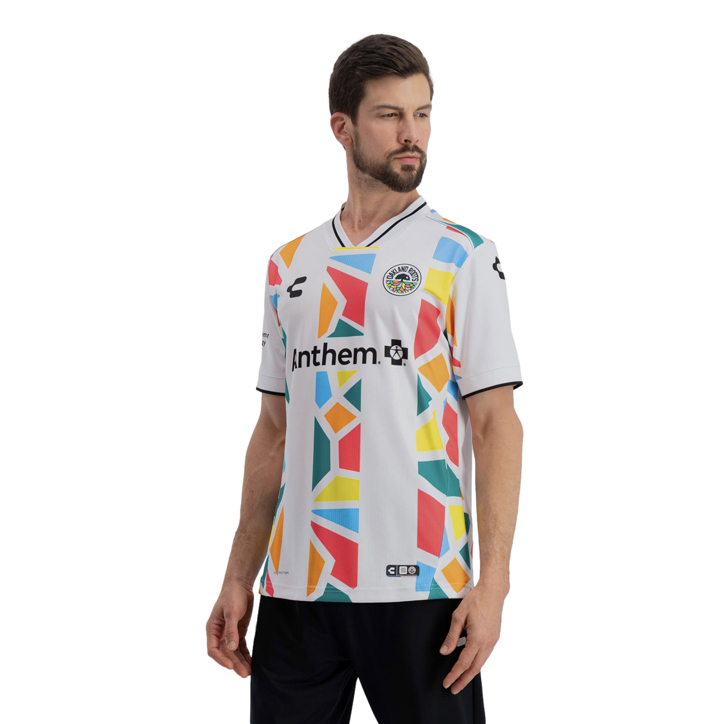 A man models a white Oakland Roots soccer jersey featuring a vibrant, multi-colored geometric mosaic pattern in shades of red, yellow, orange, blue, and green. The word "Anthem" is printed in black across the chest, and the jersey includes a black-and-white striped V-neck collar. He is posed against a solid black background.