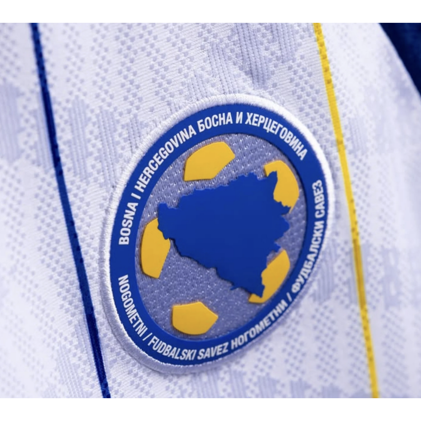 Close-up of the Bosnia and Herzegovina football association crest on a white jersey. The circular blue badge shows a country map within a stylized yellow soccer ball.