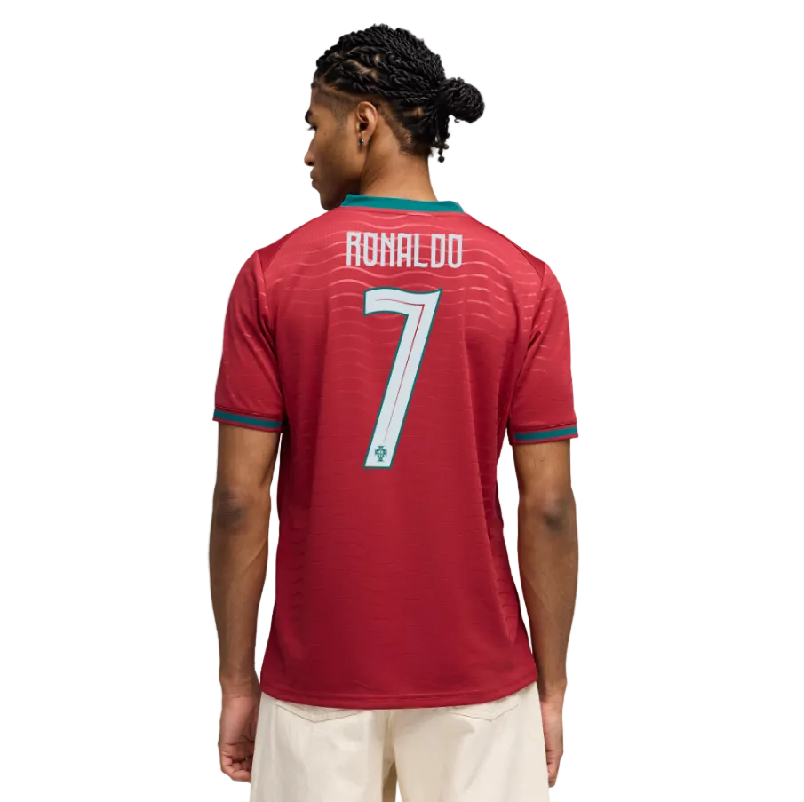 Back of a man wearing a red Portugal soccer jersey with "RONALDO 7" printed on the back. He has braided hair and wears light-colored shorts against a black background.