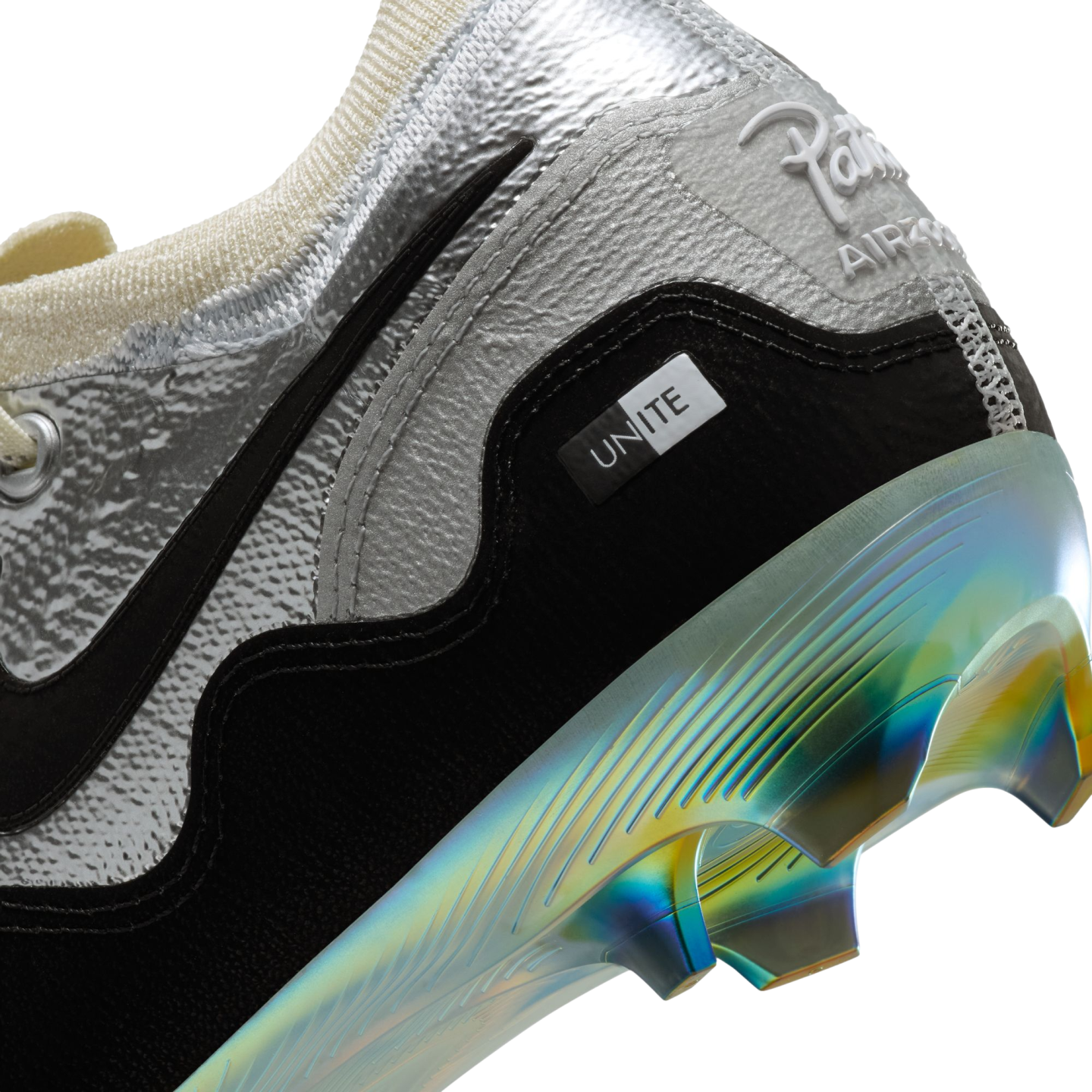 A close-up view of the heel of a silver and black Patta x Nike soccer cleat. The shoe features a textured metallic silver upper, a black suede-like base with a small white "UNITE" tag, and white "Patta AIR ZOOM" branding on the heel. The sole plate and studs are made of an iridescent material reflecting shades of blue, yellow, and green.
