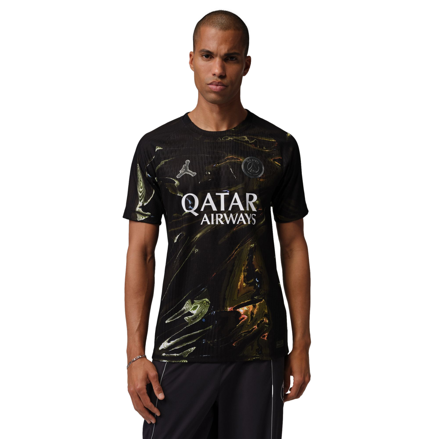 A man models a black Paris Saint-Germain soccer jersey featuring a gold and green marbled pattern. The jersey displays a silver Jordan Jumpman logo, the club crest, and "QATAR AIRWAYS" in white across the chest. He is standing against a solid black background wearing dark athletic pants with white piping.