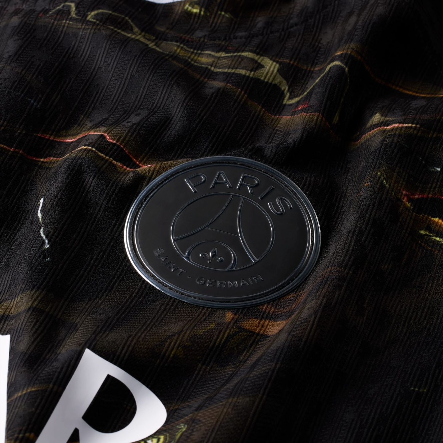 A close-up photograph of a matte black Paris Saint-Germain (PSG) football club badge. The circular badge, featuring raised lettering and the team's iconic Eiffel Tower and fleur-de-lis logo, is set against a dark, ribbed fabric jersey with marbled gold and pink patterns.