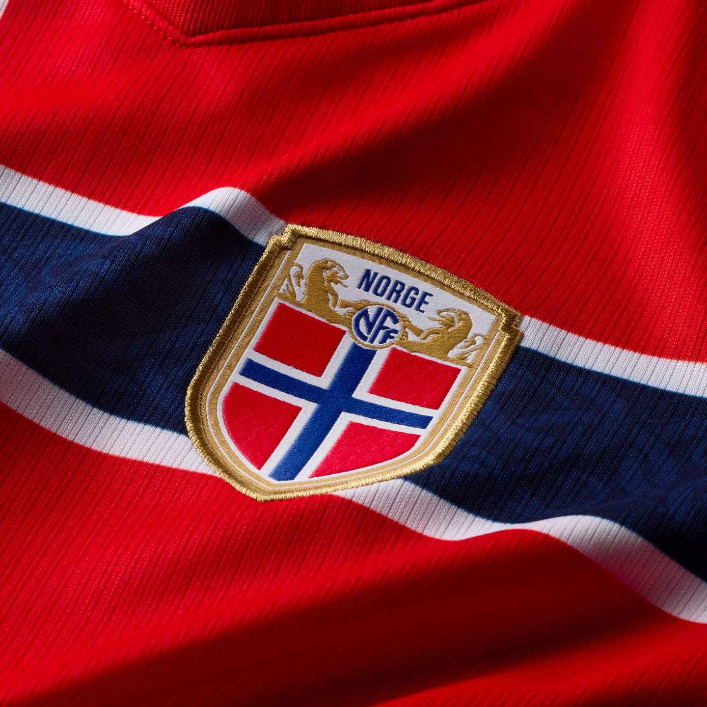 A close-up of a red Norway national team football jersey. A horizontal navy blue stripe with white borders runs across the fabric. Centered on the stripe is the Norwegian Football Federation (NFF) crest, featuring a gold-bordered shield with the Norwegian flag, two gold lions, and the word "NORGE" in blue. The fabric has a visible vertical ribbed texture.
