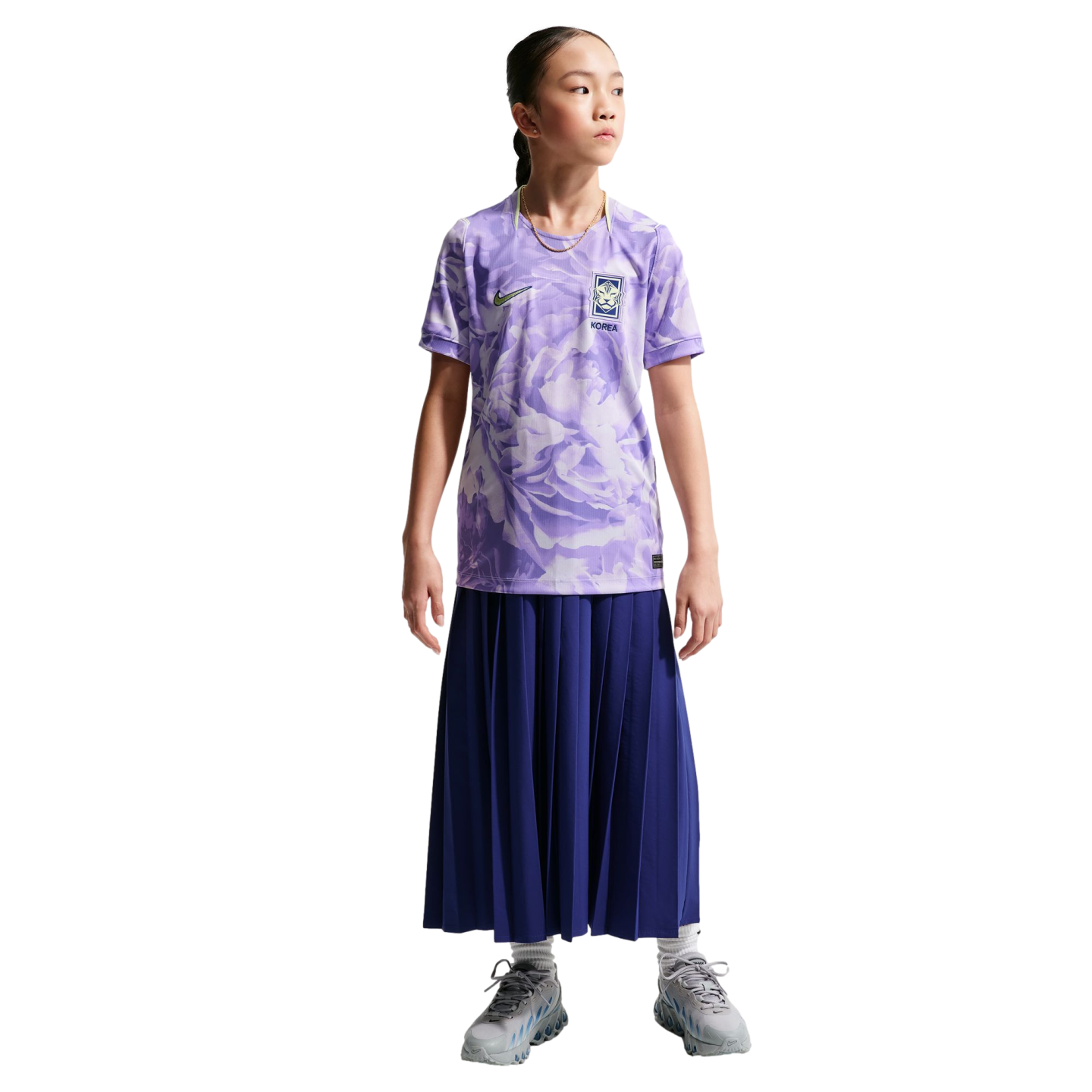 A young girl in a purple floral Nike South Korea soccer jersey, long navy pleated skirt, and gray sneakers, standing against a black background.