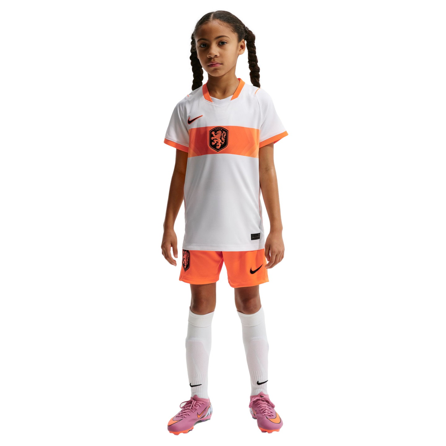 A young girl with braided pigtails stands against a black background, wearing a white and orange Nike Netherlands national team soccer kit, white socks, and pink cleats.