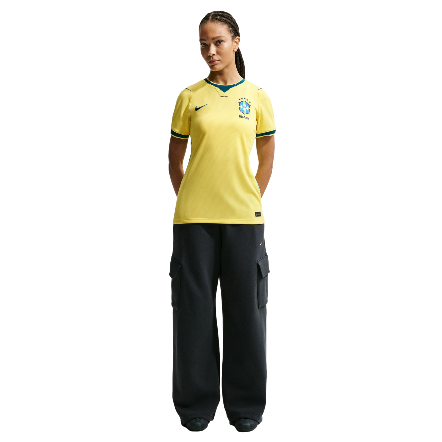 A woman with braided hair wearing a yellow Brazil national soccer team jersey and dark cargo pants, standing against a black background.