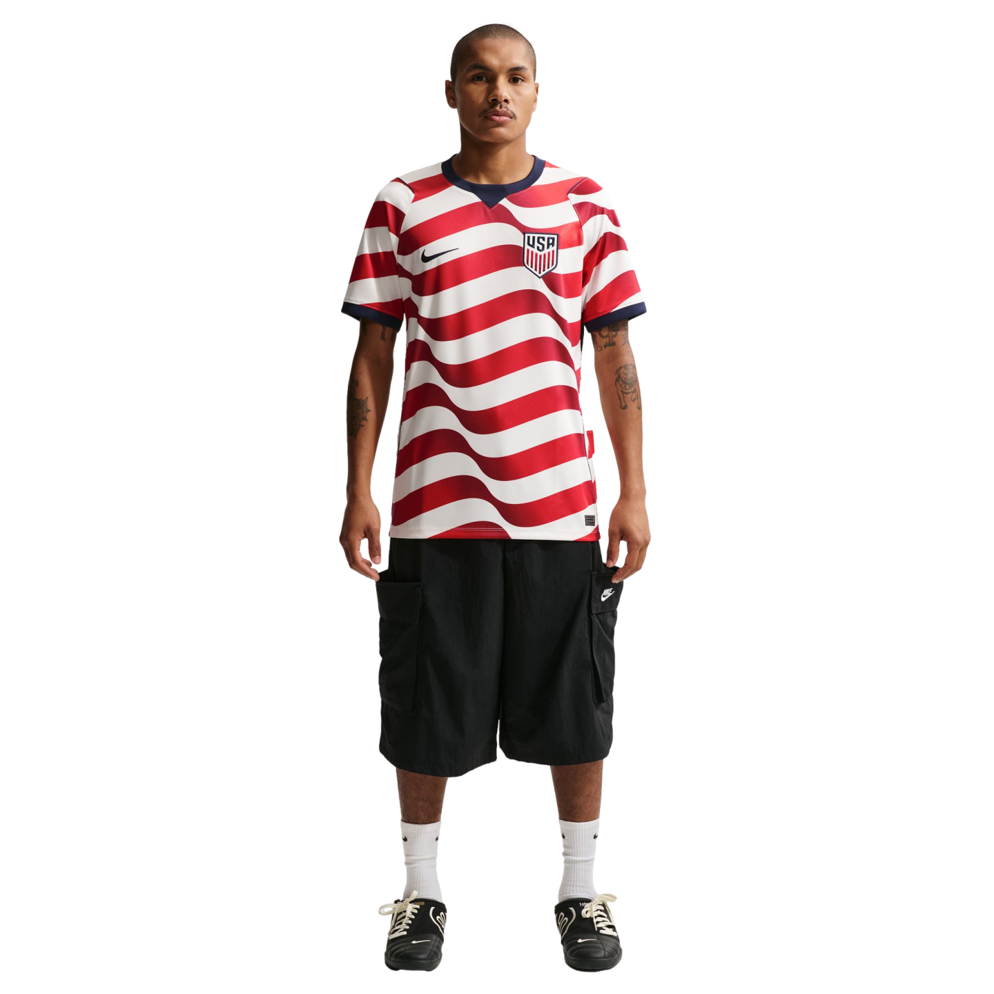 Man wearing a red and white wavy-striped USA soccer jersey, black cargo shorts, and black sneakers, standing against a black background.