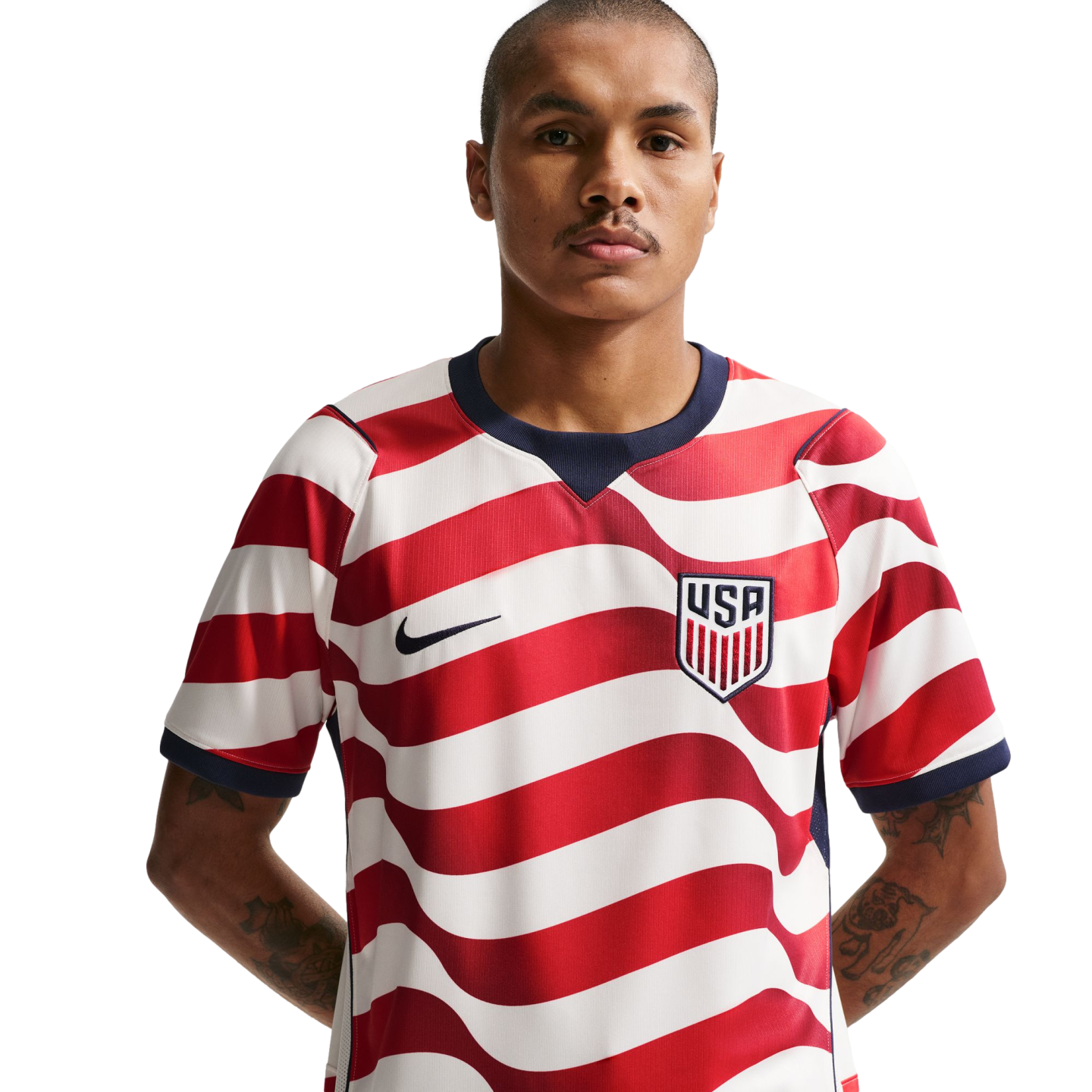 A man with a mustache in a red and white wavy-striped USA soccer jersey with a Nike logo and USA crest, against a black background. He has tattoos on his forearms.