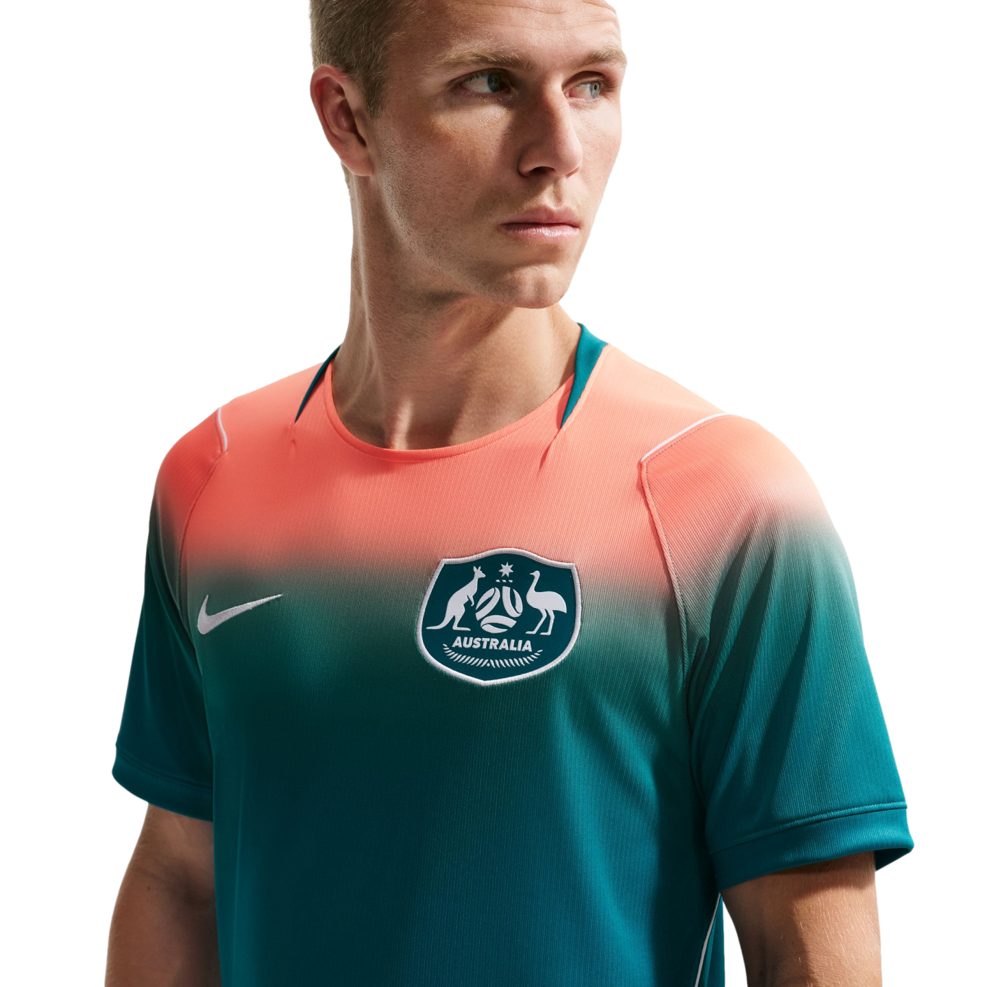 A man wearing an Australian national soccer team jersey with a salmon-to-teal color gradient, featuring the team crest and a white Nike logo. He is looking off-camera against a solid black background.