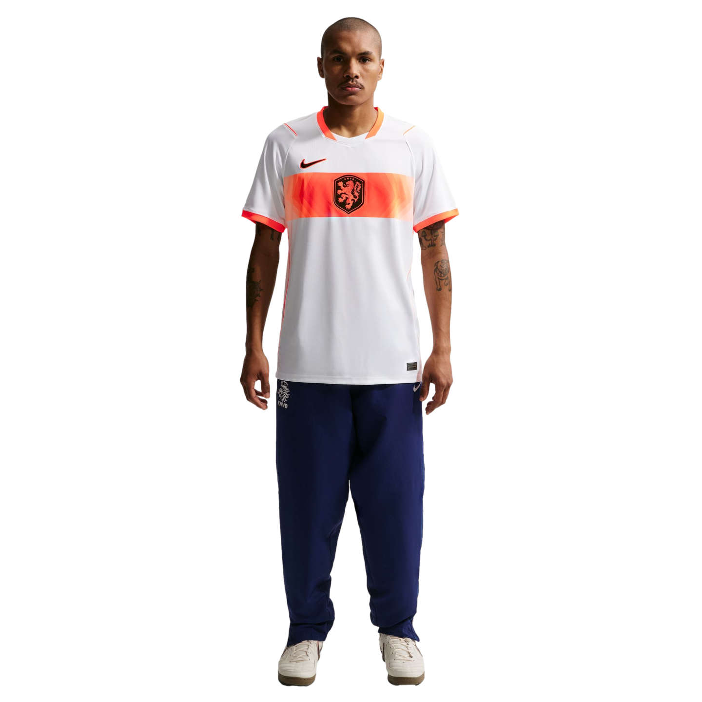 A full-length studio portrait of a man standing against a black background. He is wearing a white Nike soccer jersey with an orange horizontal band across the chest that features a black lion crest. He is also wearing navy blue tracksuit pants and off-white sneakers. He has short hair, tattoos on his arms, and is looking directly at the camera with a neutral expression.