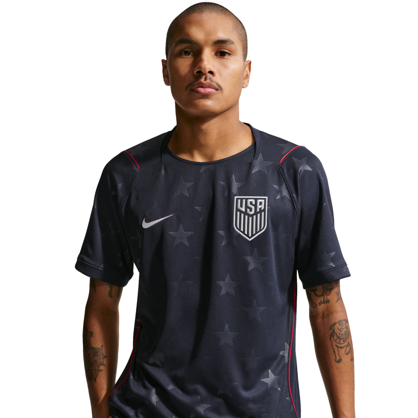 A man wearing a dark blue Nike USA soccer jersey with a subtle star pattern and red trim. He has tattoos on his arms and stands against a black background.
