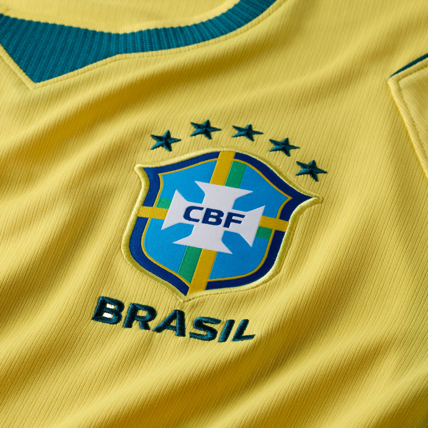 A close-up shot of a yellow Brazilian national soccer team jersey, featuring the embroidered CBF crest in the center. Above the crest are five green stars arranged in an arc, and the word "BRASIL" is embroidered in green underneath. A portion of the dark green ribbed V-neck collar is visible at the top.