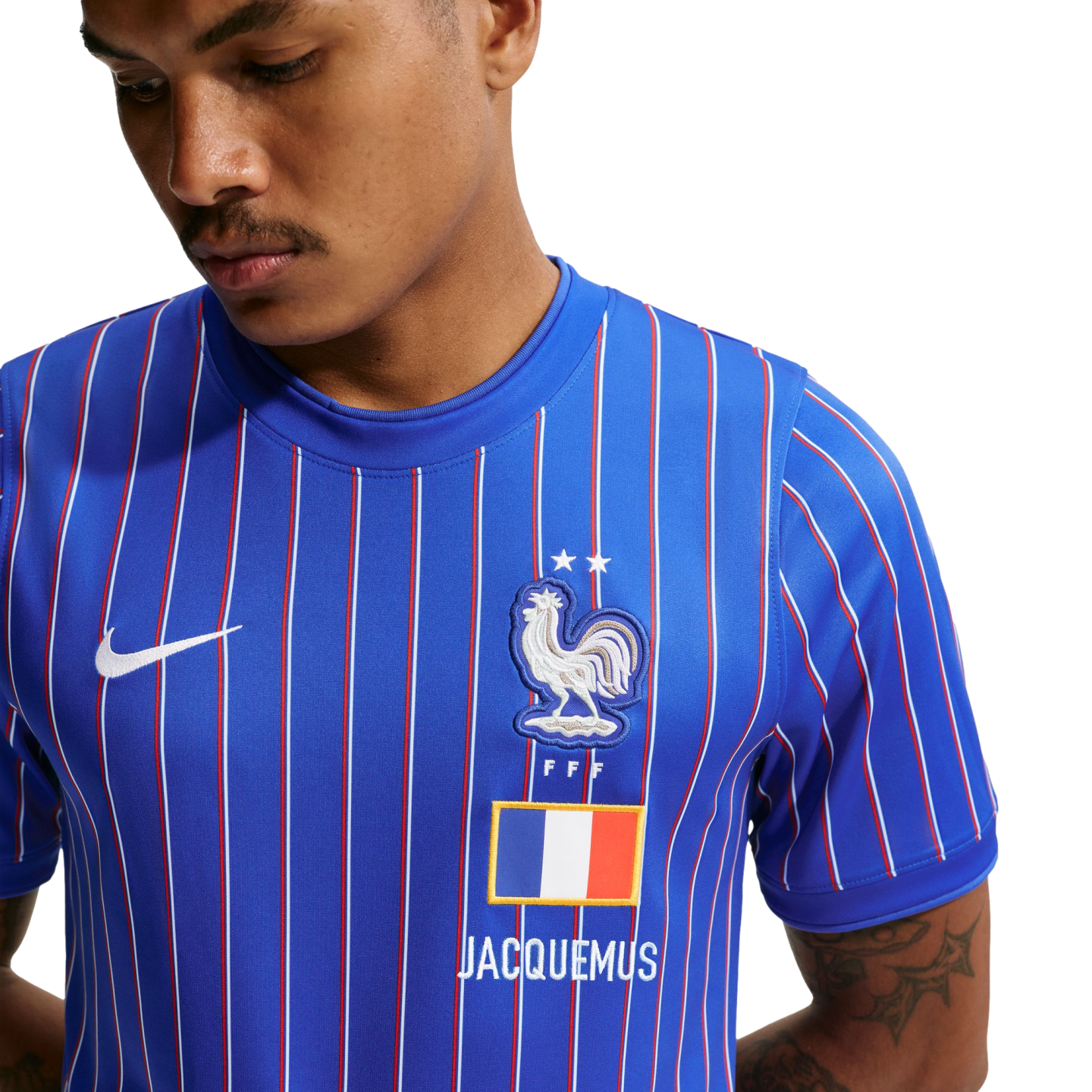 Close-up of a man wearing a blue Nike x Jacquemus France football jersey with red and white pinstripes, featuring the FFF rooster crest, two stars, and a French flag.