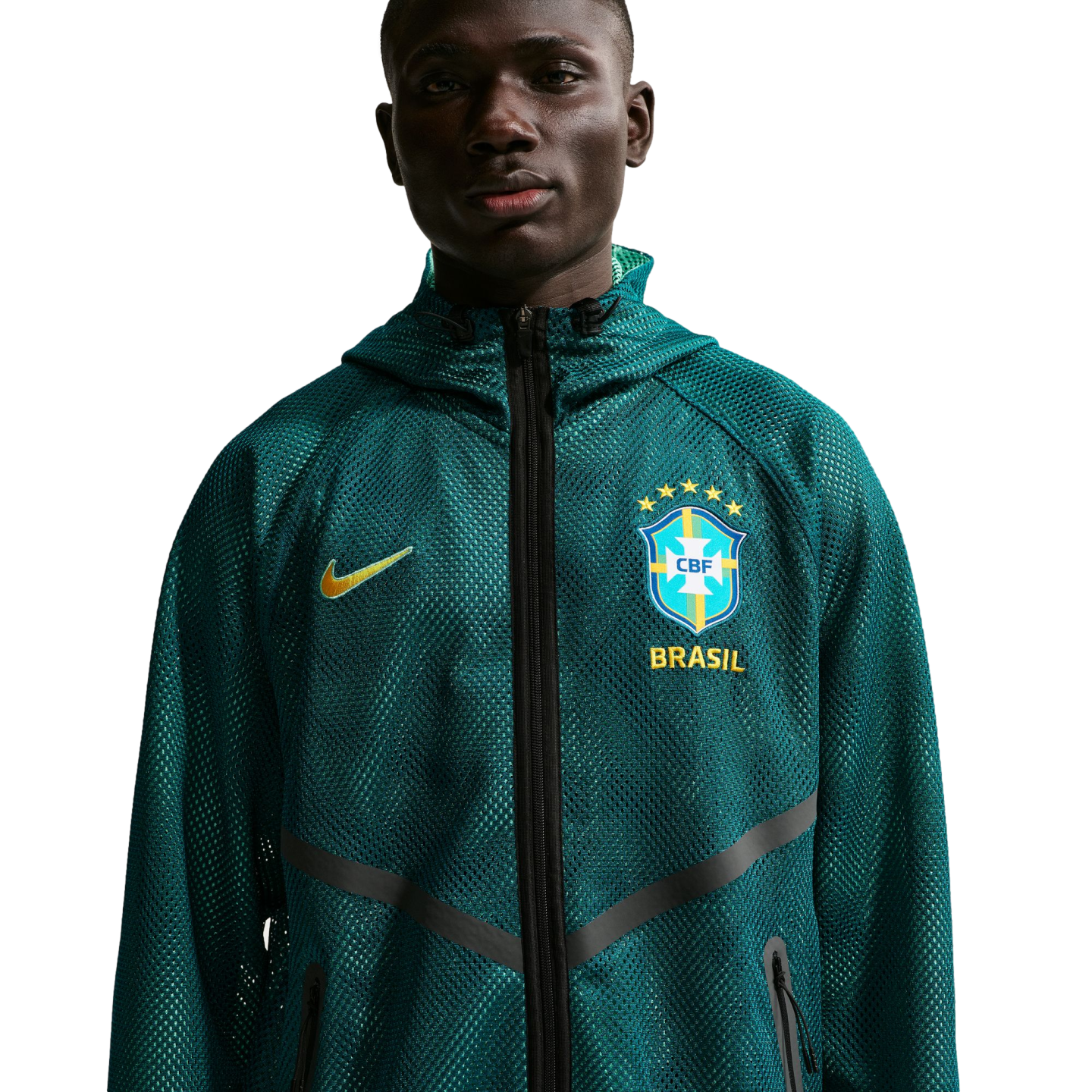 A waist-up portrait of a young Black man wearing a dark teal mesh Nike zip-up hoodie. The hoodie features a yellow Nike swoosh on one side and the Brazil national football team crest with "BRASIL" printed below it on the other. He is looking directly at the camera against a solid black background.