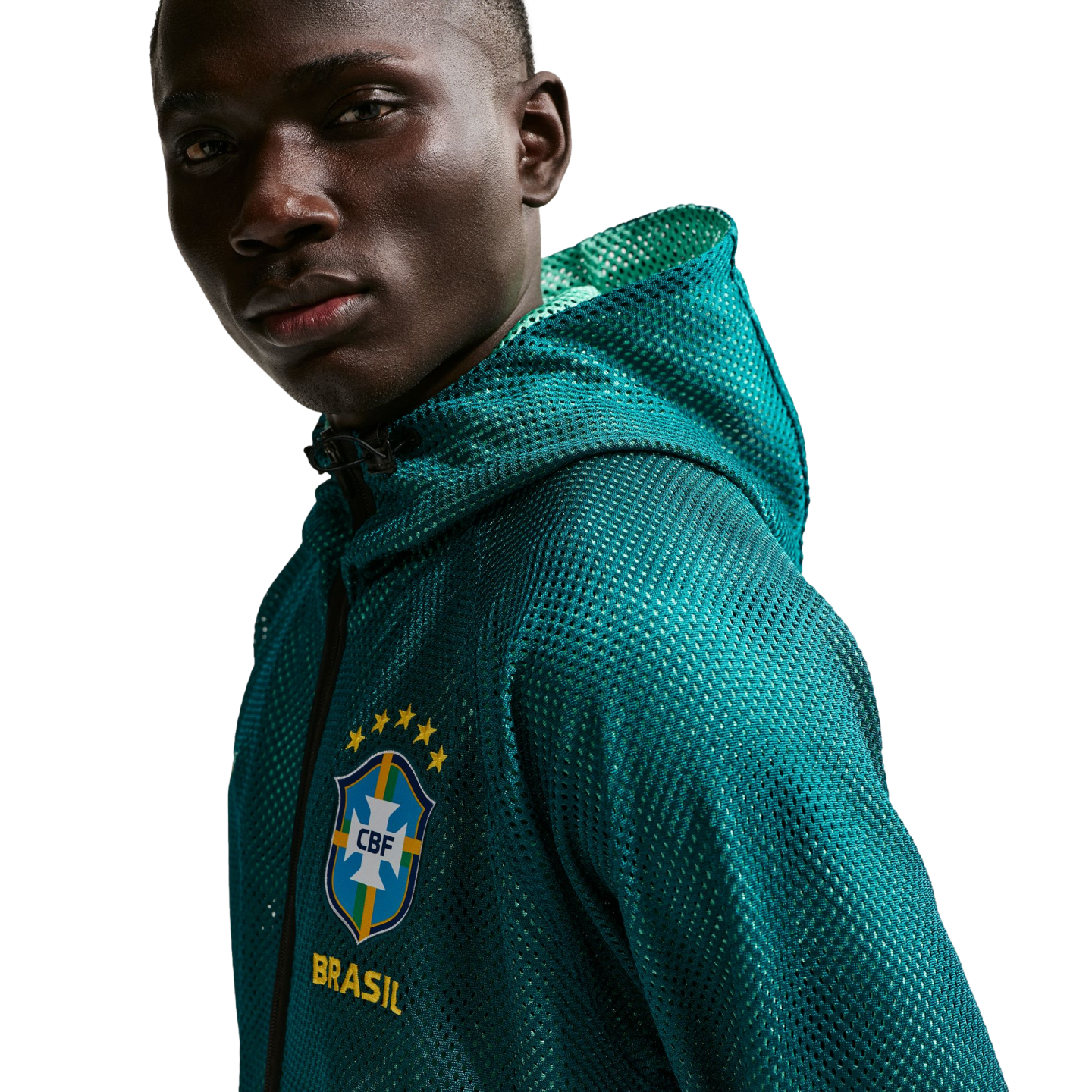 A close-up shot of a man with dark skin looking towards the camera against a solid black background. He is wearing a teal, mesh, zip-up hoodie with the Brazilian football confederation (CBF) crest on the chest, featuring five yellow stars above it and the word "BRASIL" in yellow below.
