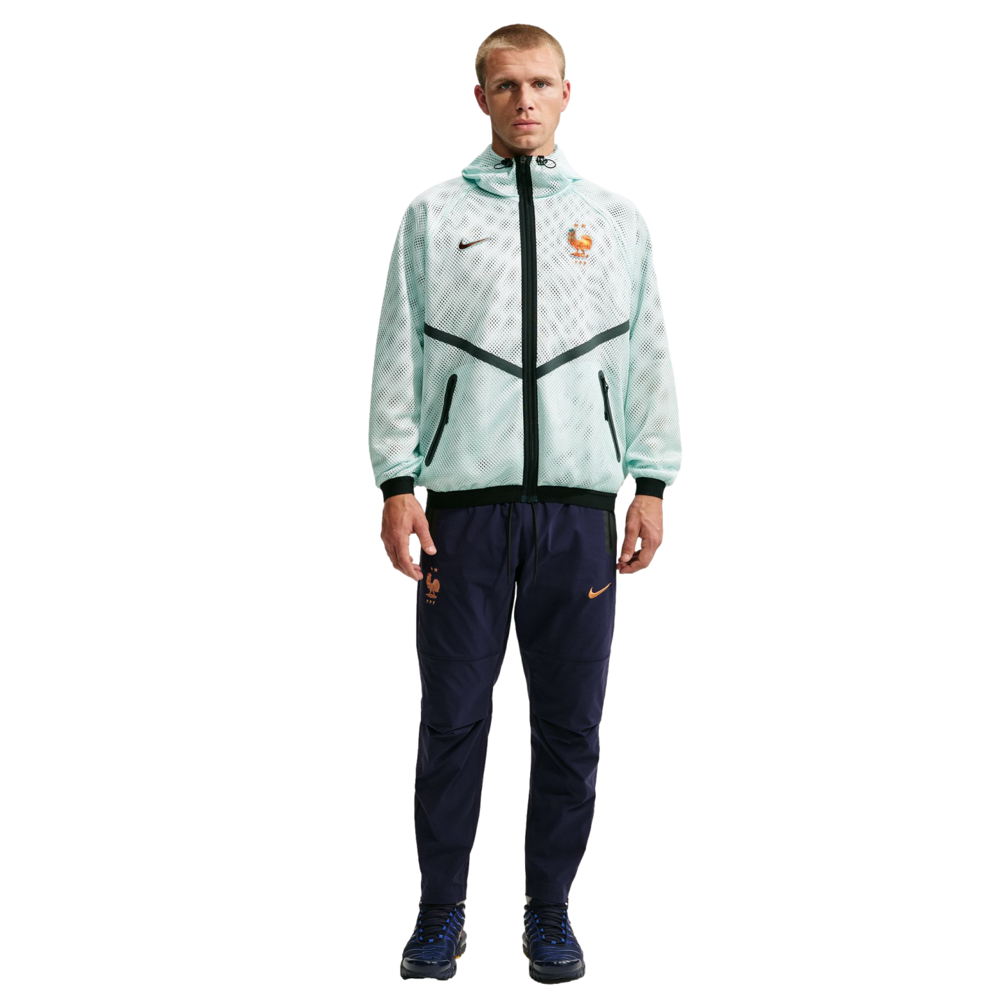 A man stands against a solid black background wearing a Nike tracksuit for the French national football team. He is wearing a light mint green, mesh-textured hooded jacket with black accents and navy blue track pants with orange logos.