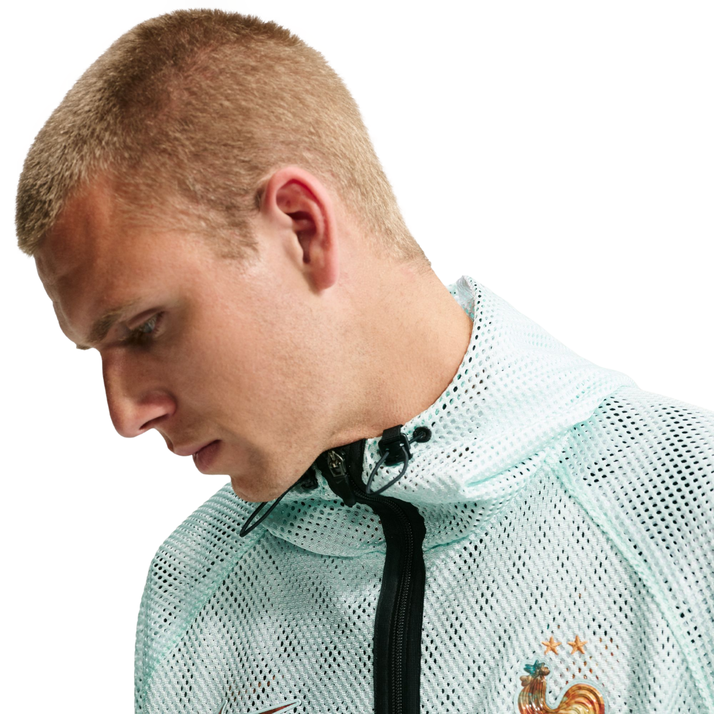A profile shot of a blond man looking down, wearing a mint green Nike mesh jacket featuring the French National Football Team crest.