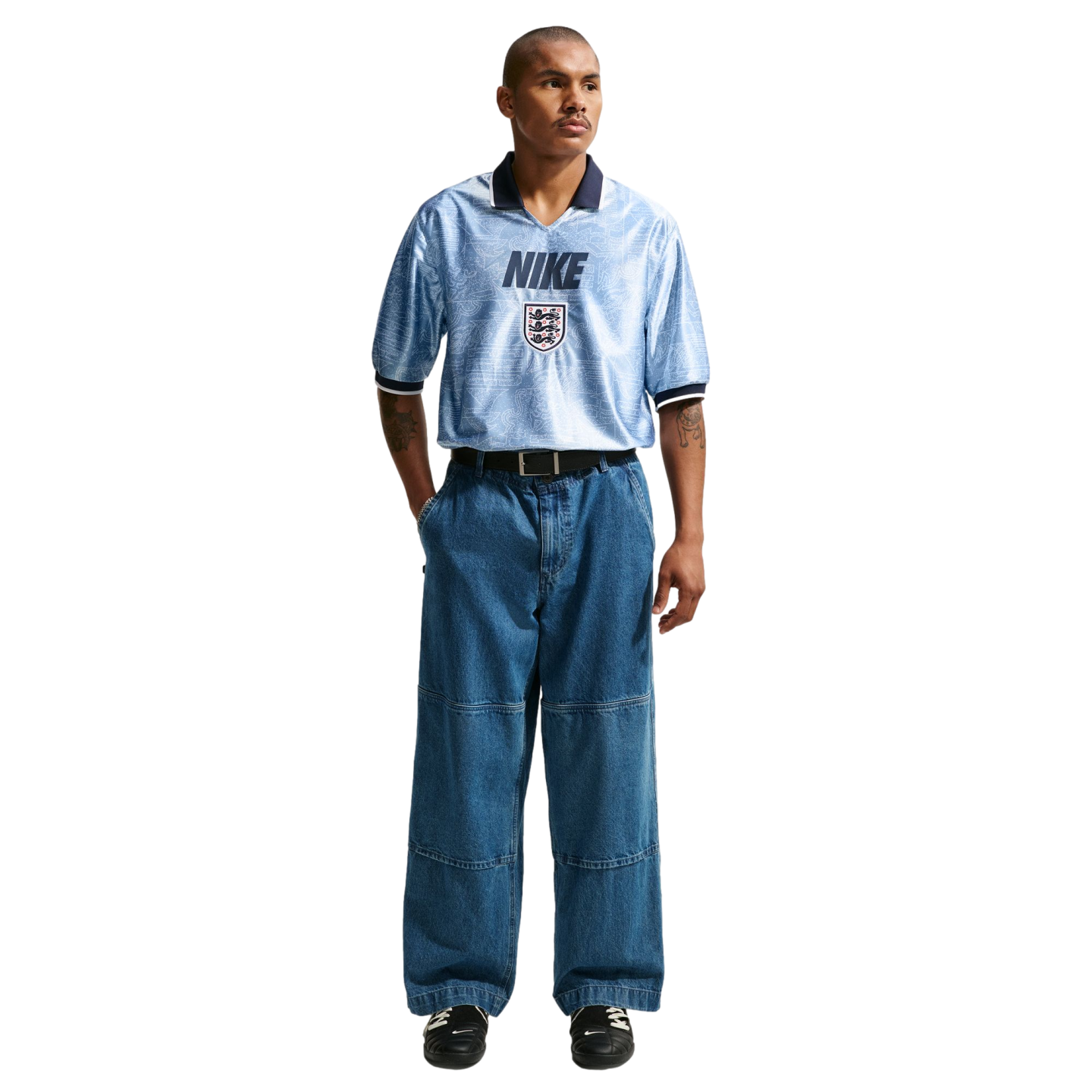 A man models a light blue Nike England jersey tucked into baggy wide-leg blue jeans and black sneakers against a plain white background.