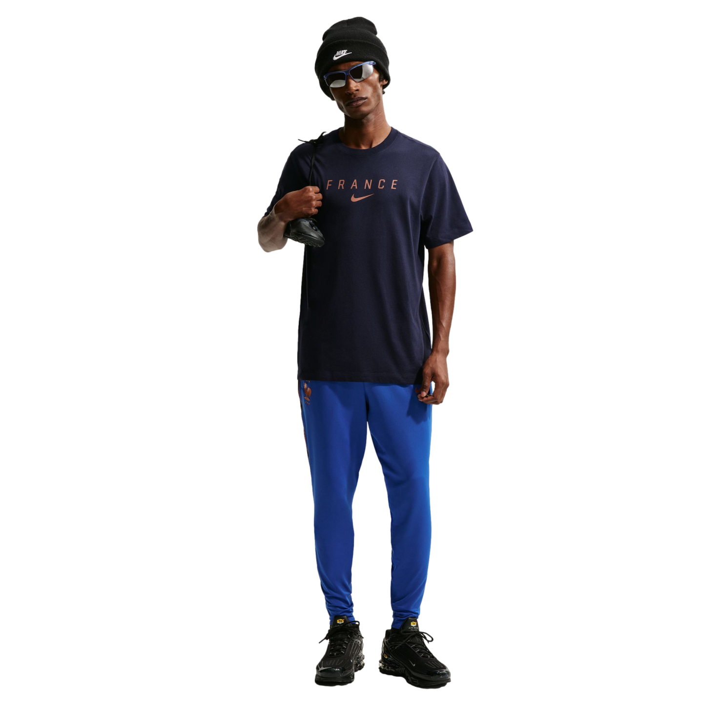 A full-body photo of a man standing against a solid black background. He is wearing a dark navy blue Nike t-shirt with "FRANCE" printed on the chest, bright blue athletic pants, black sneakers, a black Nike beanie, and blue-tinted sunglasses. He is holding a black shoe over his right shoulder.