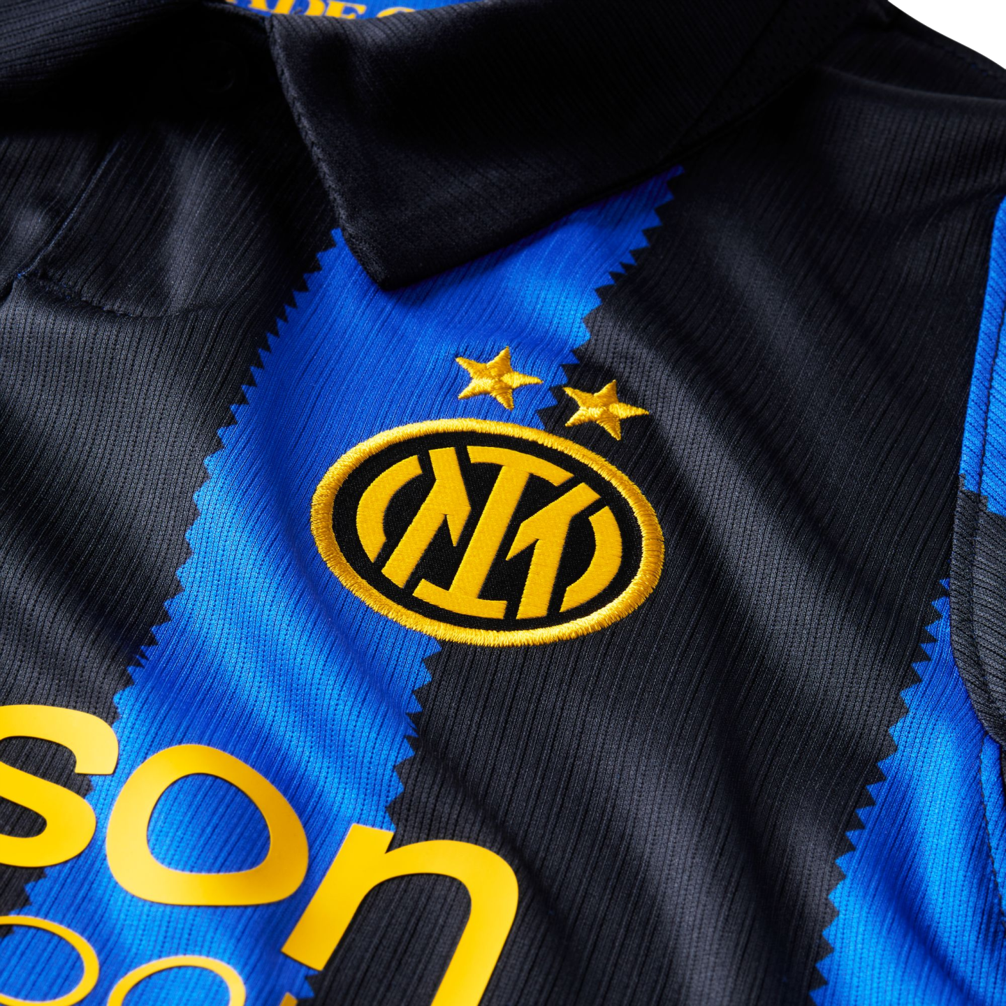 Close-up of an Inter Milan jersey with blue and black jagged stripes. It features the team's gold crest under two stars and part of a yellow sponsor logo.