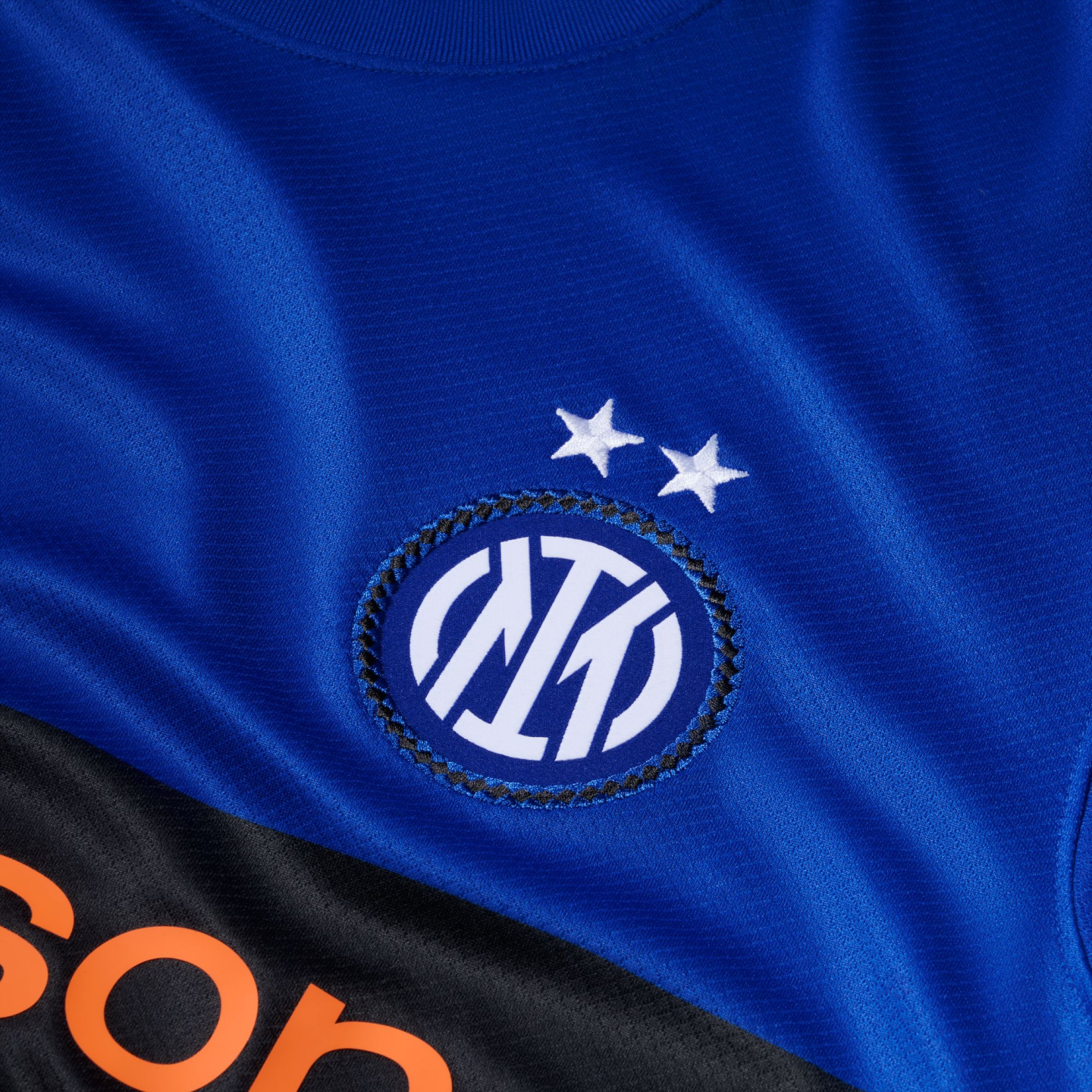 Close-up of a blue Inter Milan soccer jersey with the club crest and two white stars above it. A black section with orange lettering is at the bottom.