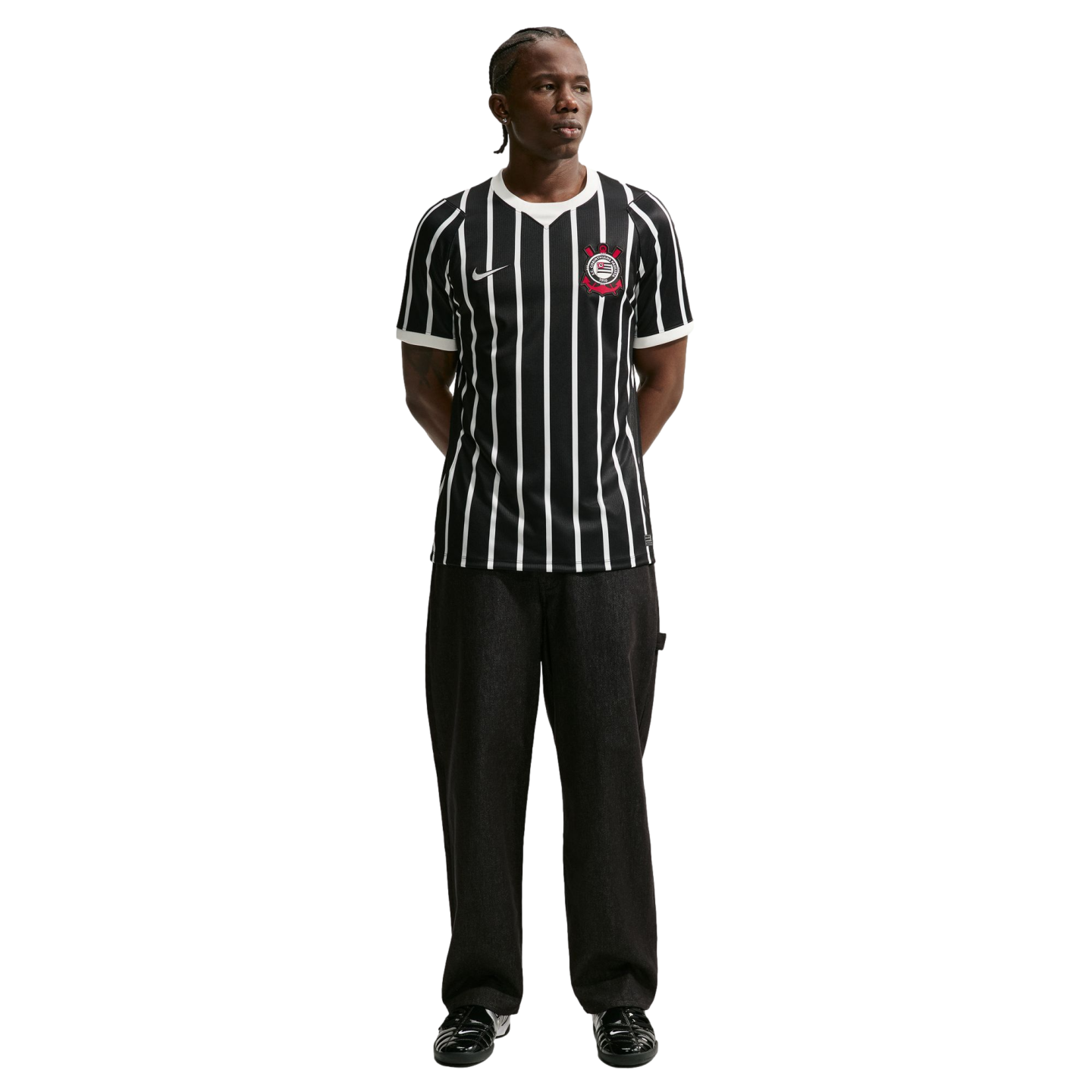 Full-length shot of a man in a black and white striped Corinthians soccer jersey, black pants, and black sneakers, standing against a solid black background.