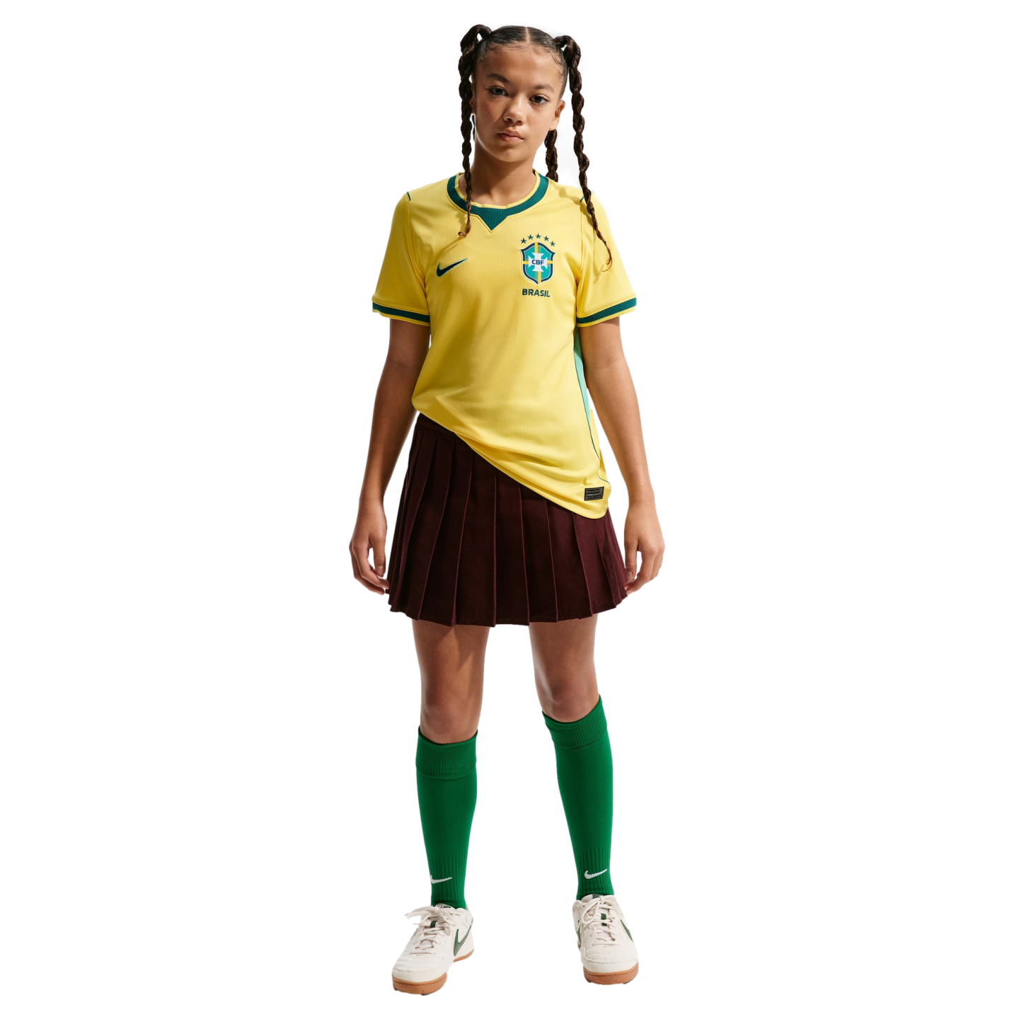 A young girl wearing a yellow Brazil soccer jersey, a maroon pleated skirt, green socks, and white sneakers, standing against a black background.