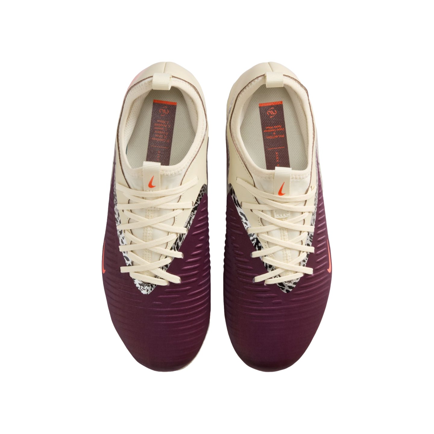 A top-down view of a pair of Nike football boots against a black background. The shoes feature a maroon lower section with horizontal ridges and a cream upper section. A black-and-white patterned trim lines the lace area, and small orange Nike swooshes are visible on the tongues and sides.