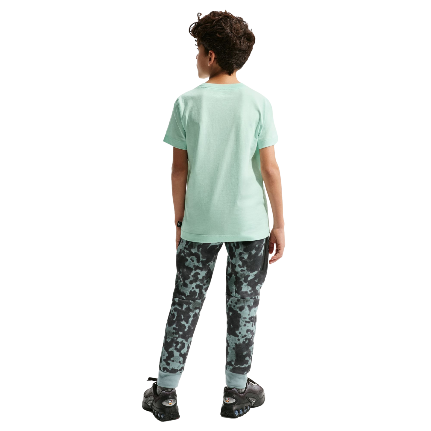 Back view of a young boy with curly brown hair standing against a black background. He is wearing a light green short-sleeved t-shirt, camouflage-patterned joggers, and black athletic shoes.