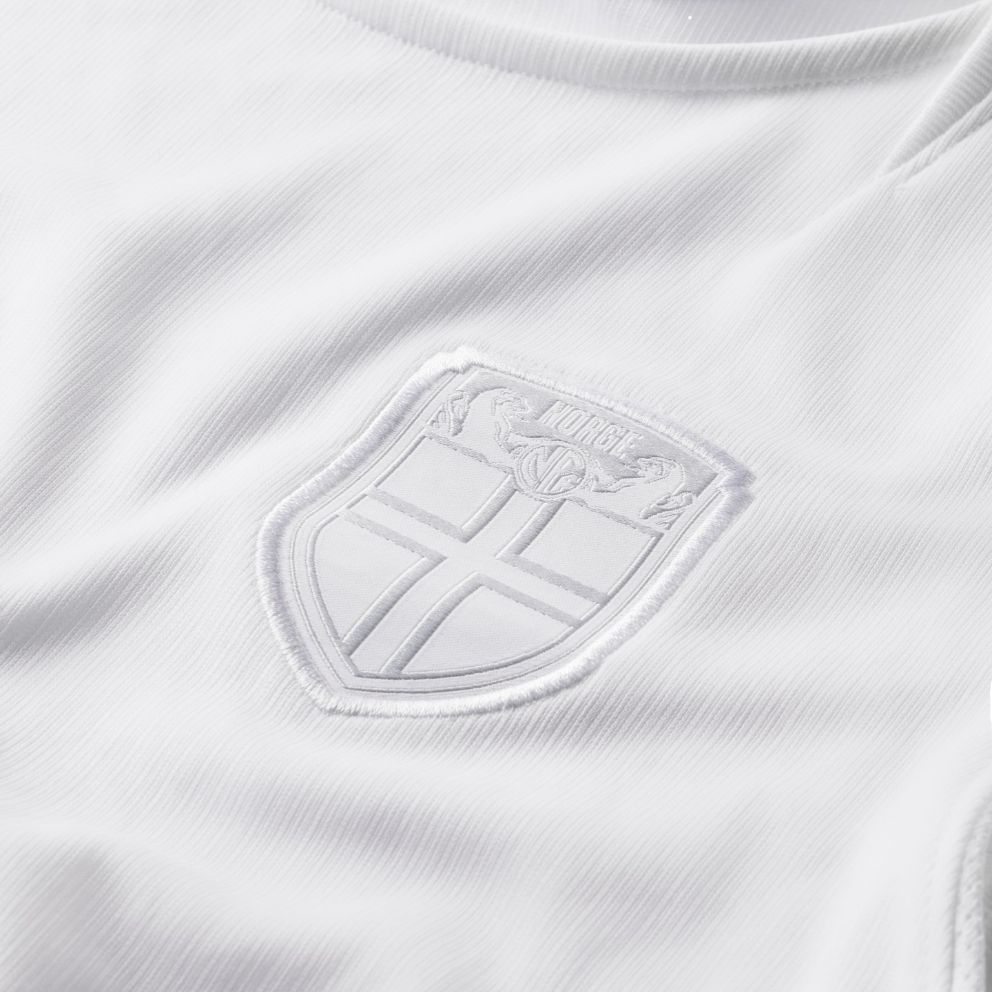 Close-up of a white textured jersey with a tonal, white-on-white embroidered crest. The shield-shaped badge features a cross and the word "NORGE" at the top.