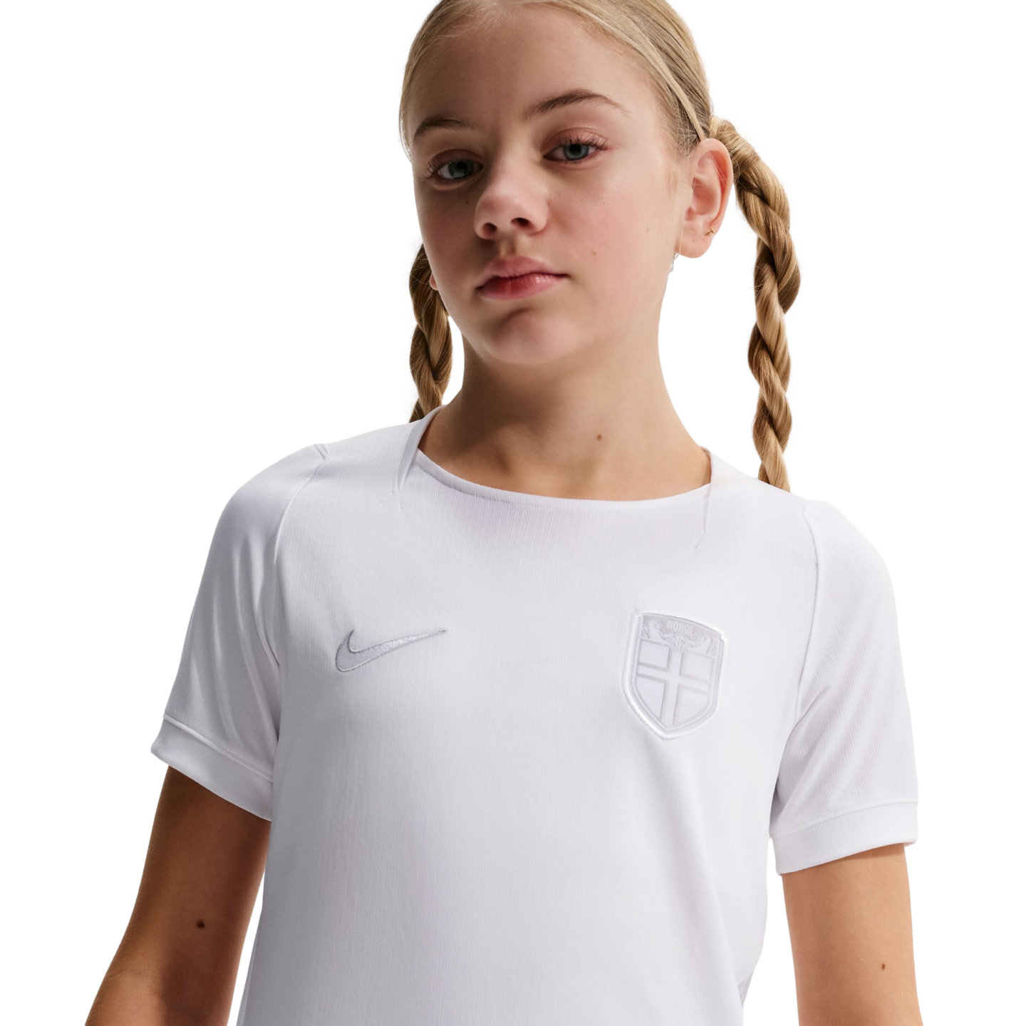Young girl with blonde pigtail braids wearing a white Nike athletic shirt with a shield crest, posed against a solid black background.