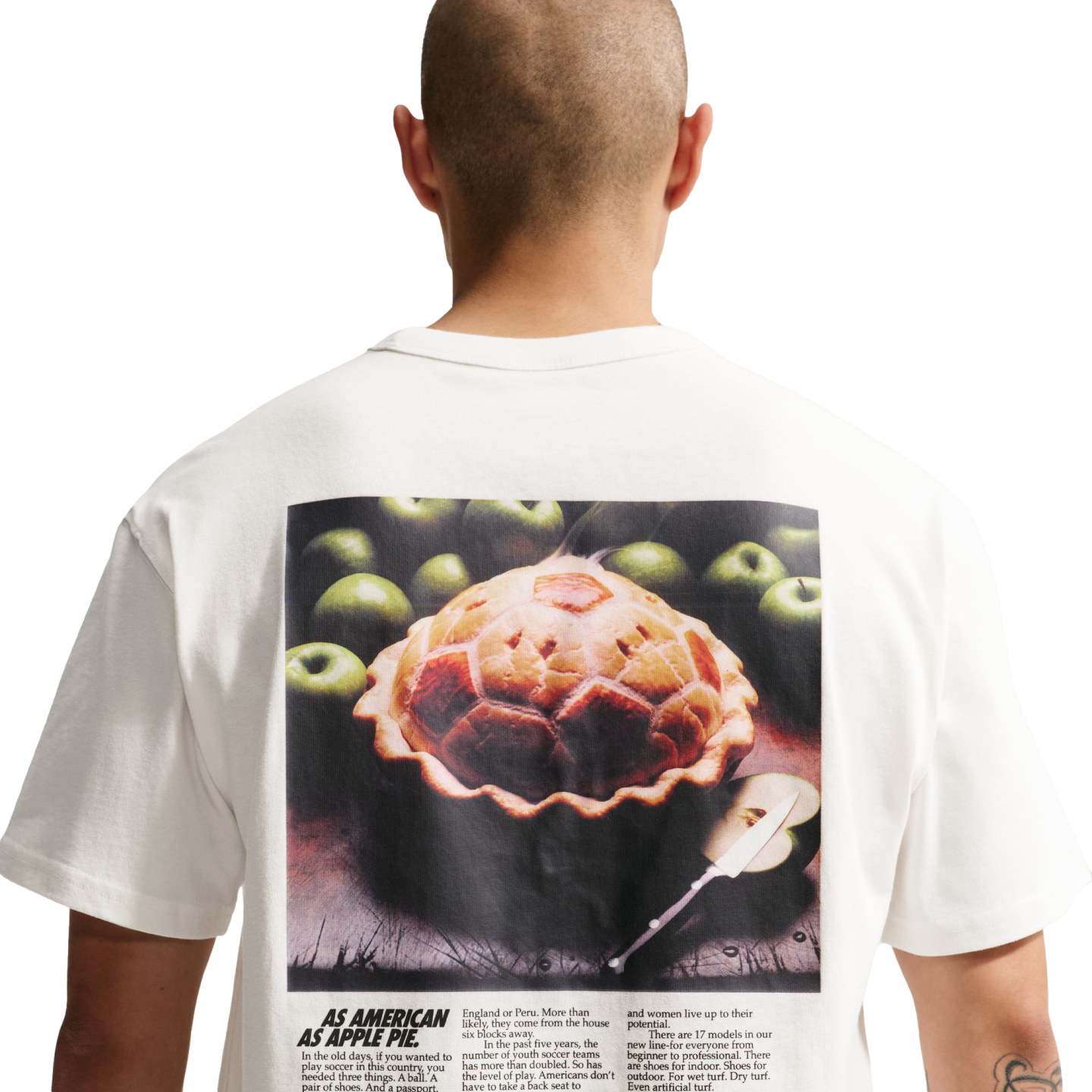 A back view of a person with a shaved head wearing a white t-shirt against a black background. The shirt features a large graphic on the back resembling a vintage magazine advertisement. The main image shows a baked pie with a soccer-ball-patterned crust surrounded by green apples. Below the image, bold text reads "AS AMERICAN AS APPLE PIE," followed by columns of text about the popularity of soccer in America and a new line of soccer shoes.