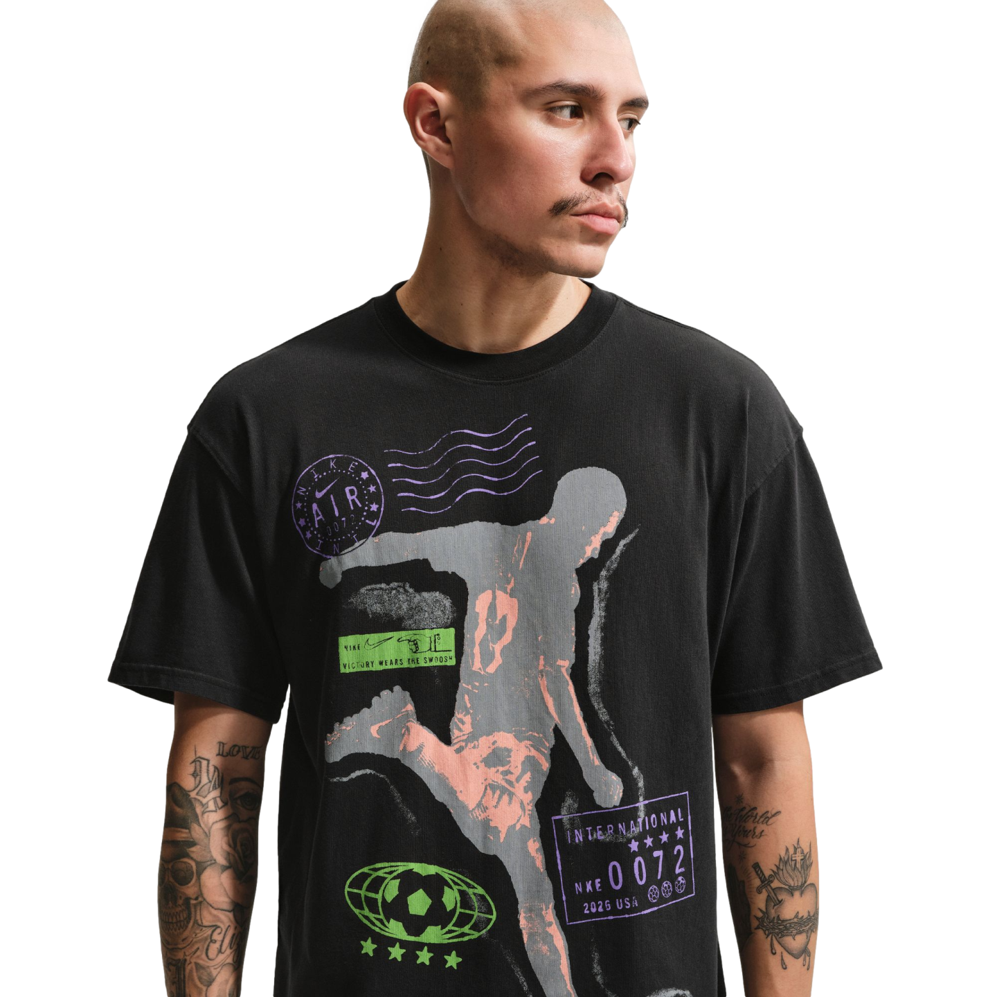 A man with a shaved head and mustache wears a black graphic t-shirt against a solid black background. The shirt features a large grey and peach-colored silhouette of an athlete in motion, surrounded by various purple and green sports-themed logos and text. Tattoos are visible on both of the man's arms.