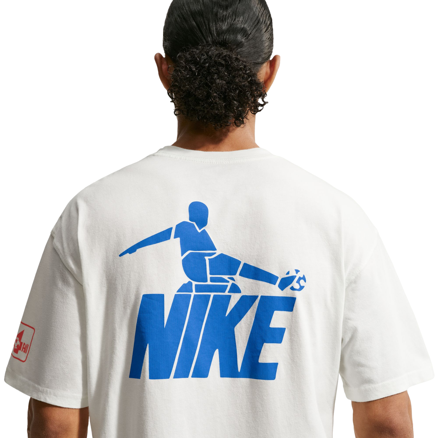A back view of a person with dark, curly hair tied in a bun, wearing a white t-shirt against a black background. The shirt features a large blue graphic on the back consisting of a stylized soccer player kicking a ball, positioned above the word "NIKE" in bold block letters.