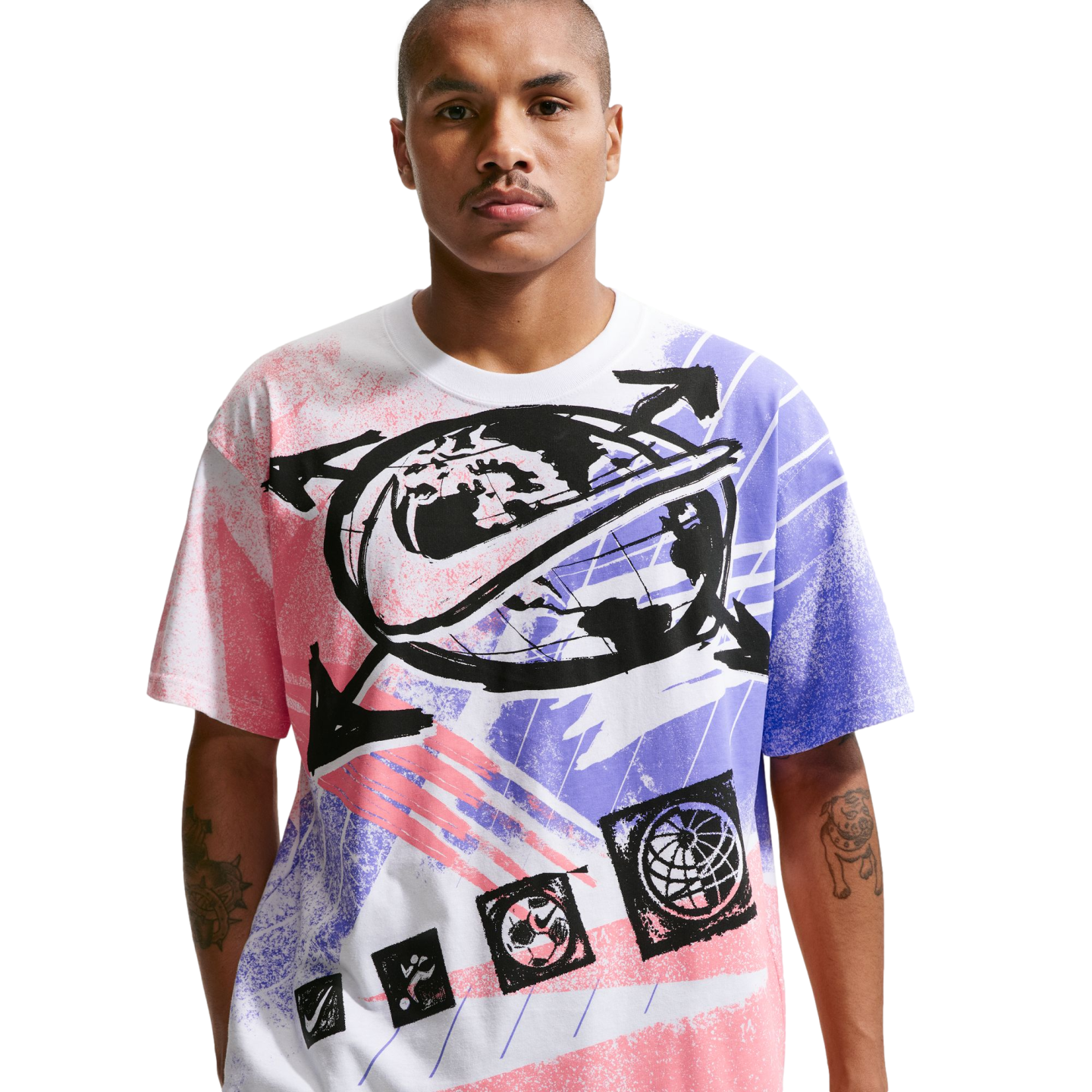 A man with short hair and a mustache stands against a solid black background, wearing a white t-shirt with a large, colorful Nike graphic. The graphic includes a central globe design with a black swoosh, surrounded by arrows and spray-paint style patterns in pink and purple. Small icons, including another globe and a Nike swoosh, are featured at the bottom of the design. He has tattoos visible on his forearms.