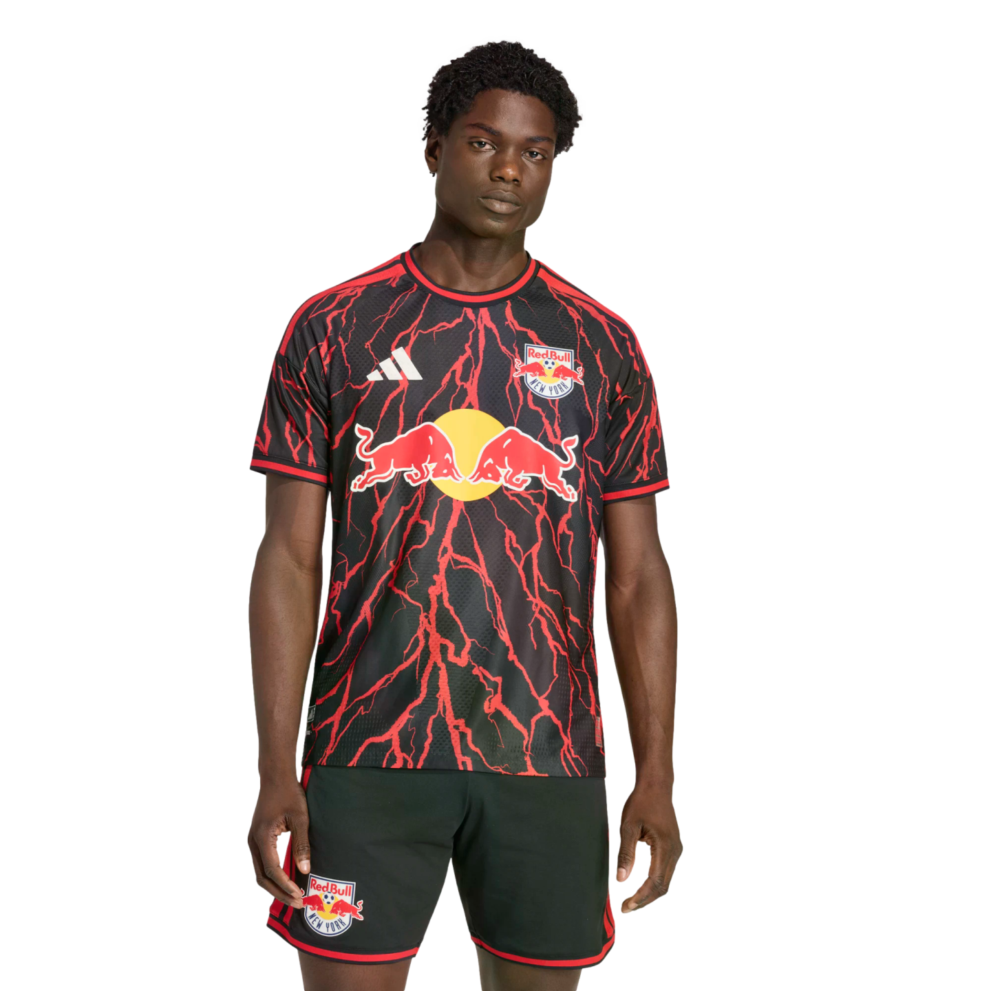 A man models a black Red Bull New York soccer jersey with a red lightning bolt pattern and matching black shorts against a black background.