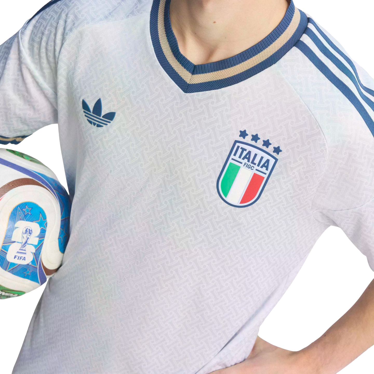 Close-up of a person in a light blue Adidas Italy football jersey with the FIGC crest, holding a FIFA soccer ball against a black background.
