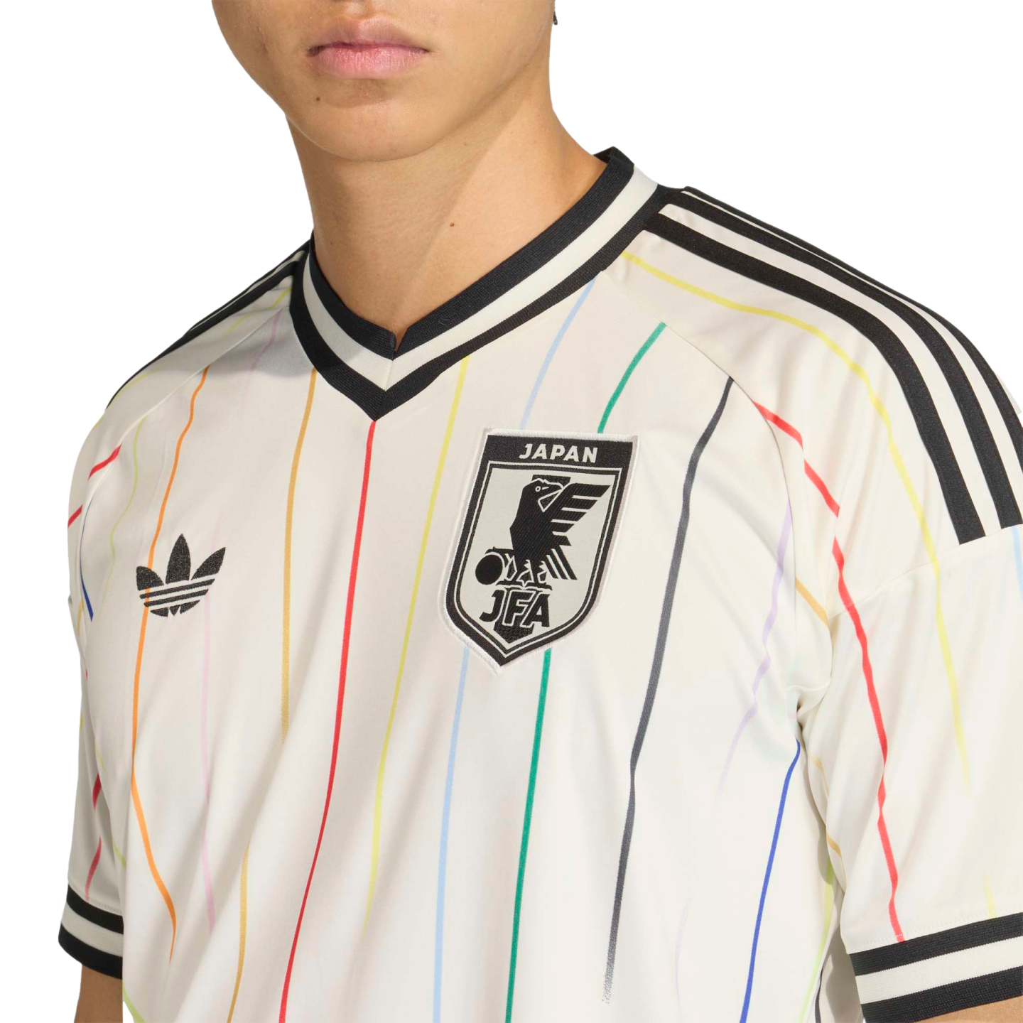 A person wearing a white Adidas Japan football jersey featuring thin vertical rainbow stripes, the JFA crest, a black trefoil logo, and black and white striped trim.