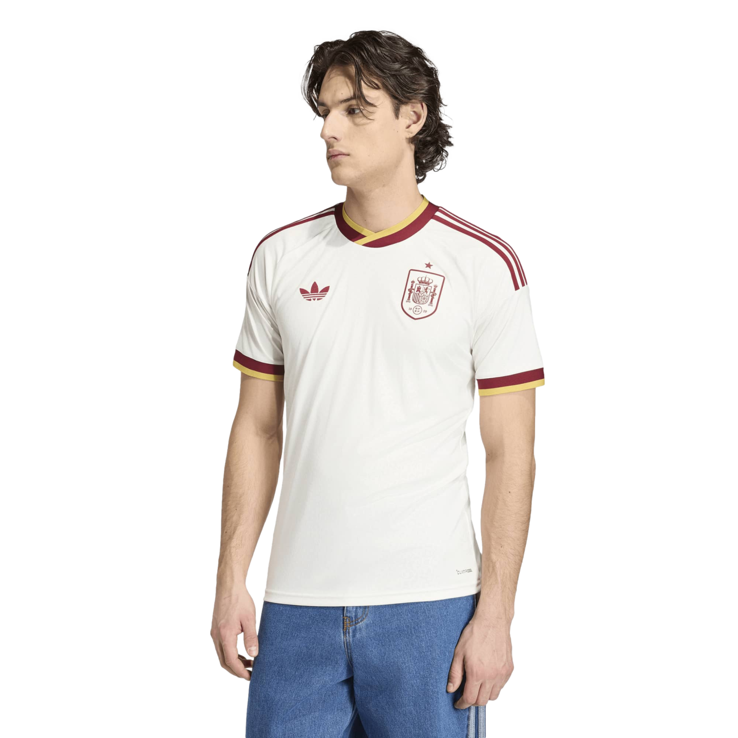 A man modeling a white Spain national football team jersey with red and yellow trim, paired with blue jeans, against a solid black background.