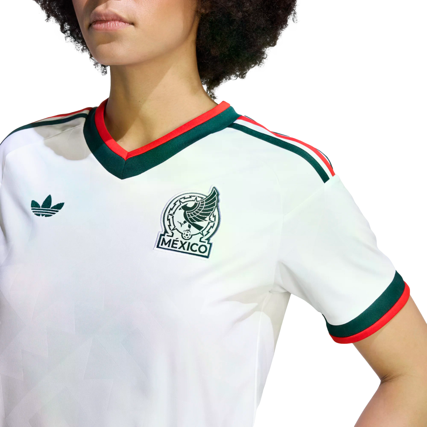 A close-up shot of a person from the chest up, wearing a white Mexico national team soccer jersey by Adidas. The jersey features a green and red V-neck collar, green and red stripes on the shoulders, and matching cuffs on the short sleeves. The Mexico national team crest is on the left chest, and a green Adidas Trefoil logo is on the right. The white fabric has a subtle, tonal geometric pattern. The background is solid black.