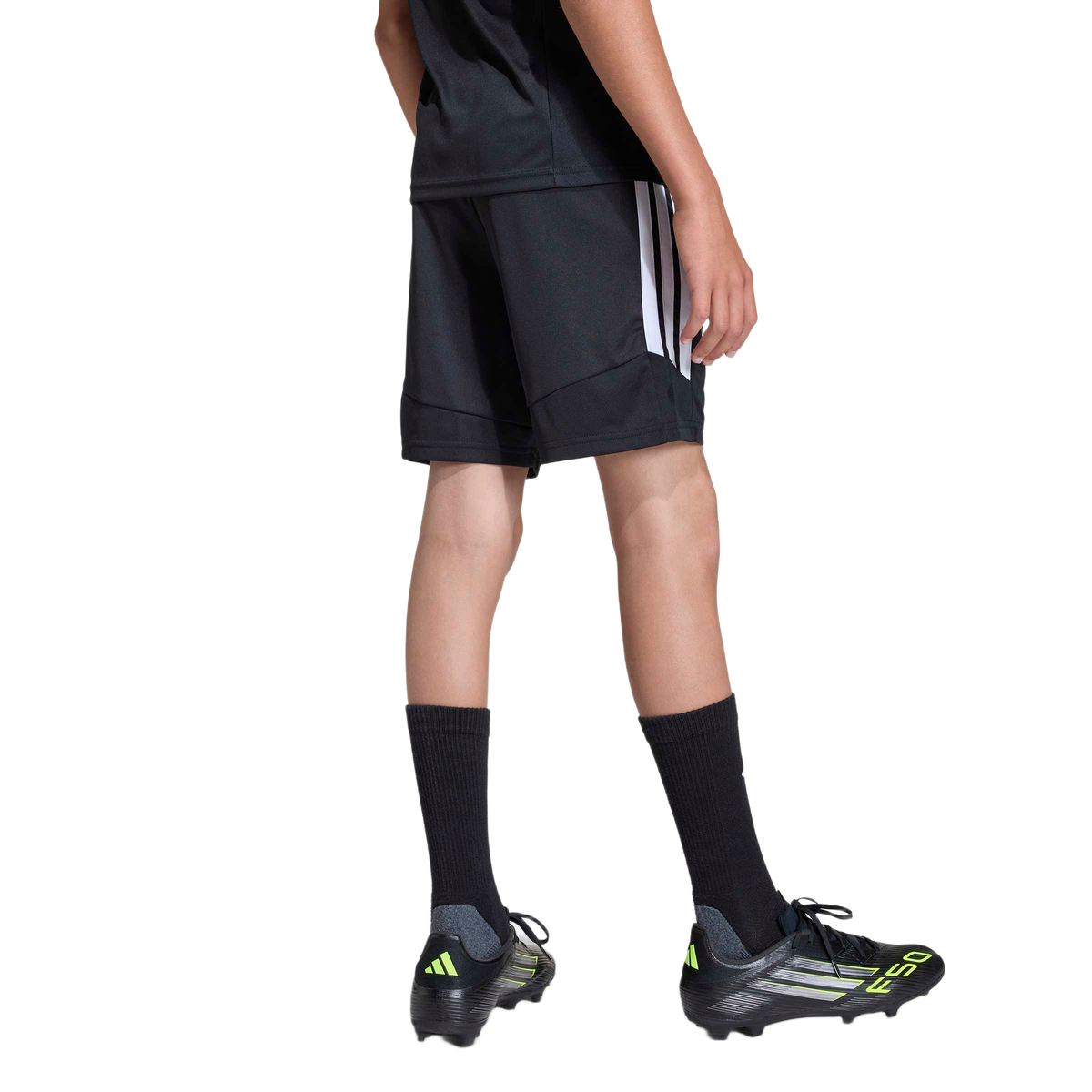 Rear view of a person wearing black Adidas athletic shorts and t-shirt, tall black socks, and black and neon yellow soccer cleats against a plain gray background.