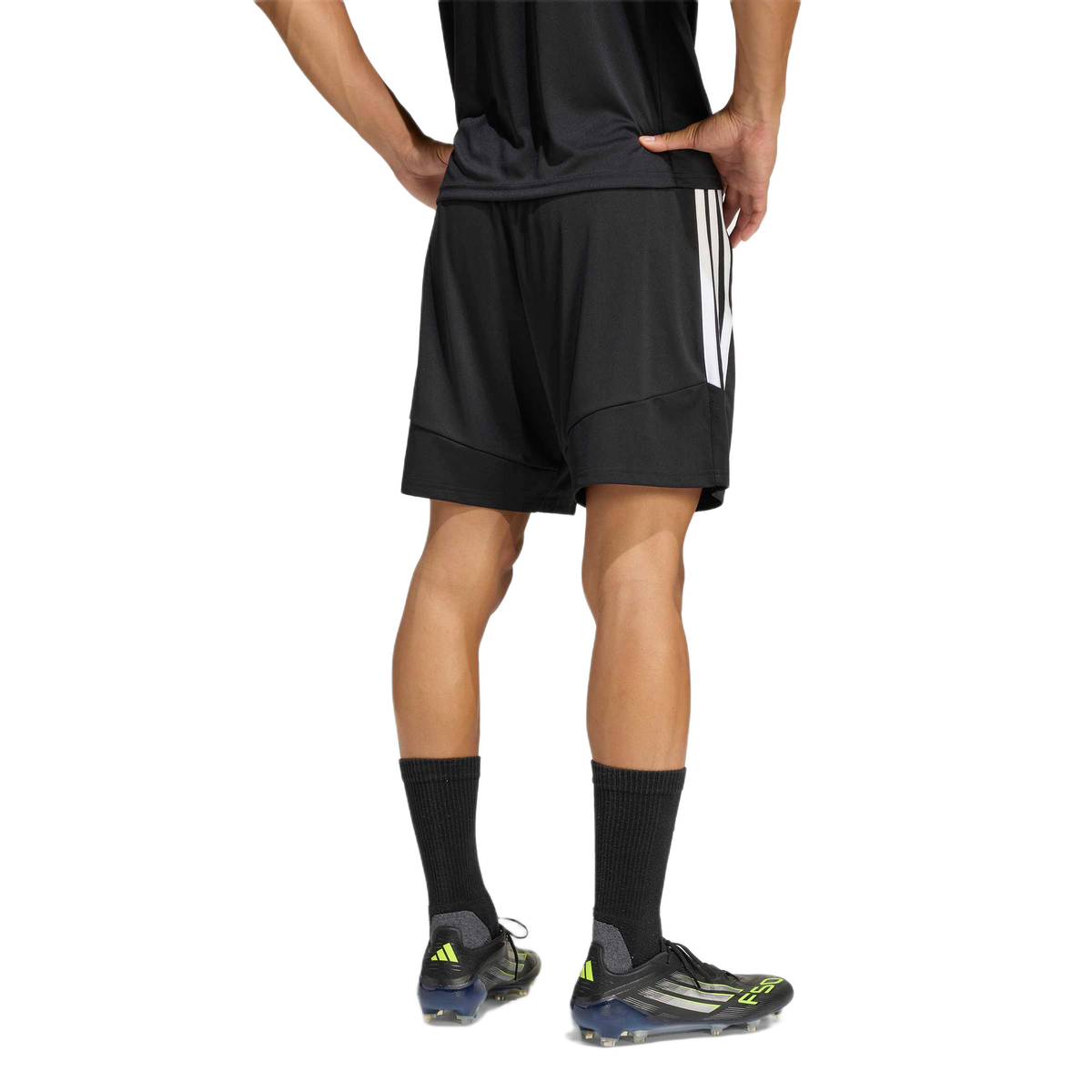 Rear view of a person wearing black Adidas athletic shorts, a black shirt, and soccer cleats against a plain white background.