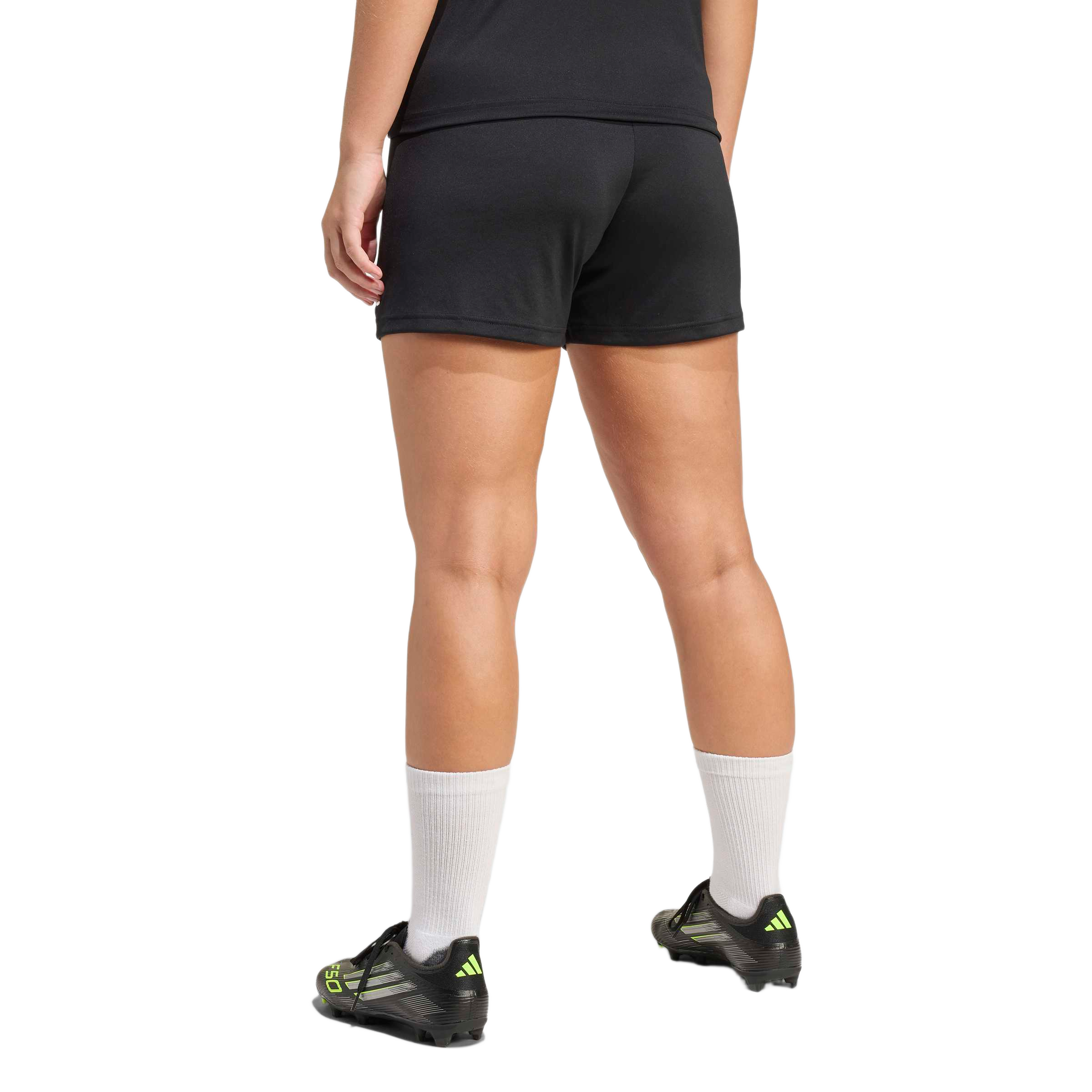 Back view of a person wearing black athletic shorts, white crew socks, and black soccer cleats with neon yellow accents against a plain grey background.