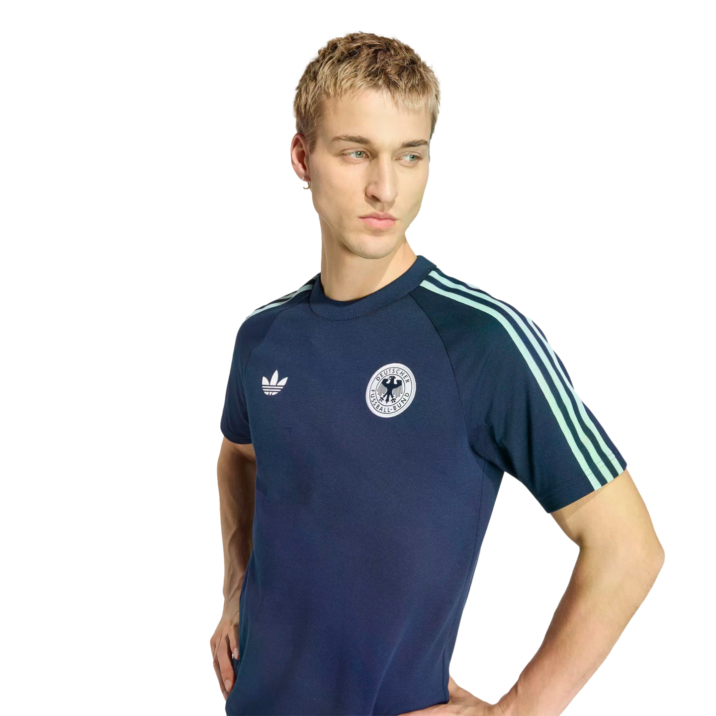 Young man with short blonde hair wearing a navy blue Adidas Germany t-shirt with mint green stripes, looking down and to the side against a black background.