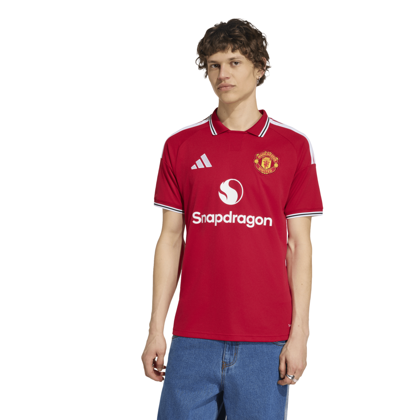A man wearing a red Manchester United home football jersey with Snapdragon sponsorship and Adidas branding, paired with blue jeans, against a plain white background.