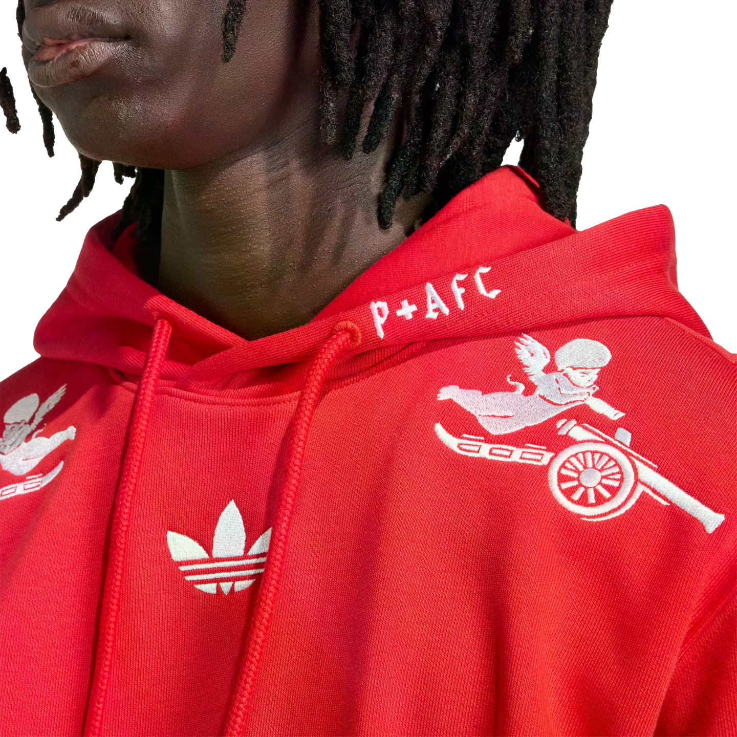 Close-up of a person in a red Adidas x Arsenal hoodie with white embroidered cherub riding a cannon, a Trefoil logo, and "P+AFC" text on the hood.